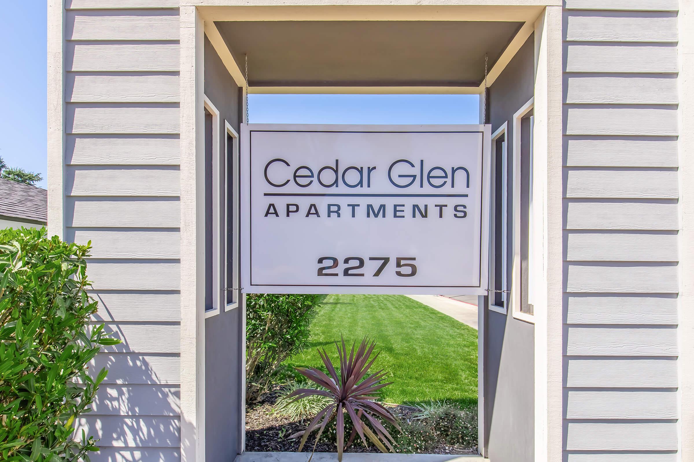 Sign for Cedar Glen Apartments, featuring the name prominently in a modern font, along with the address "2275." The sign is framed by a decorative entrance, with green shrubs and well-maintained grass visible in the background, indicative of an inviting residential complex.