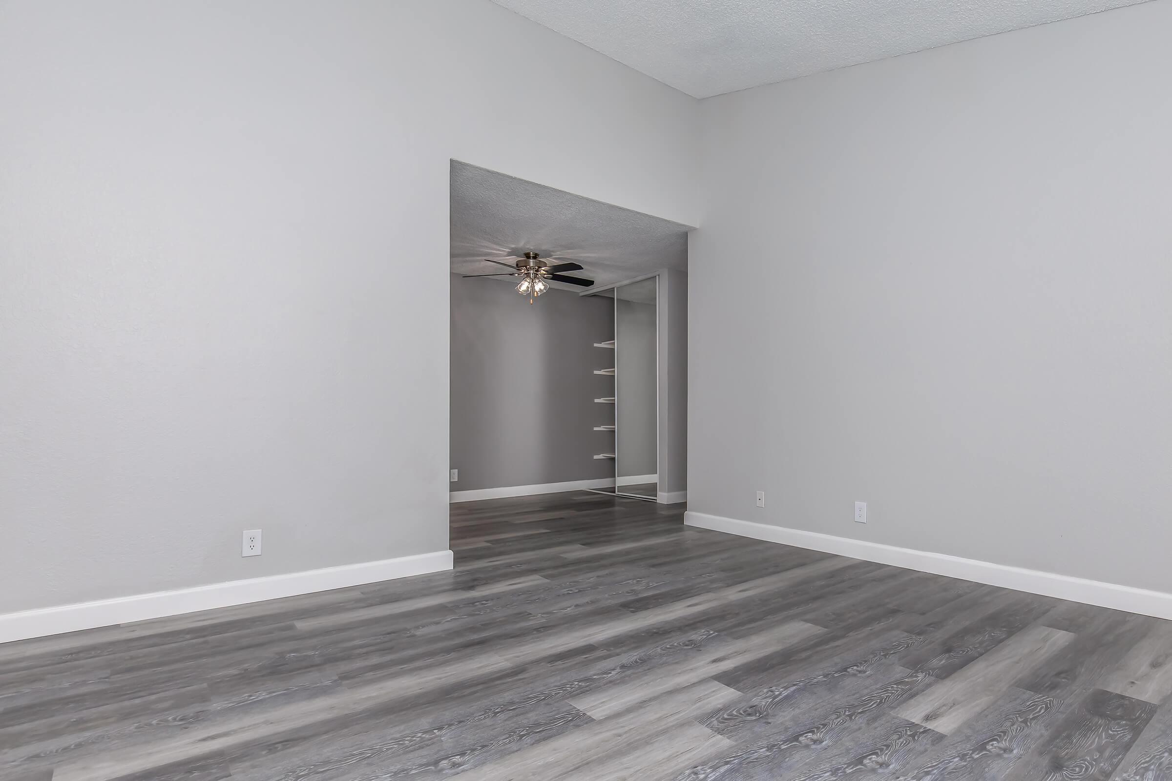 A spacious, empty room with light gray walls and sleek gray laminate flooring. Natural light streams in from a doorway leading to another area, where a ceiling fan is visible. The overall ambiance is modern and minimalistic, ideal for various furniture arrangements.