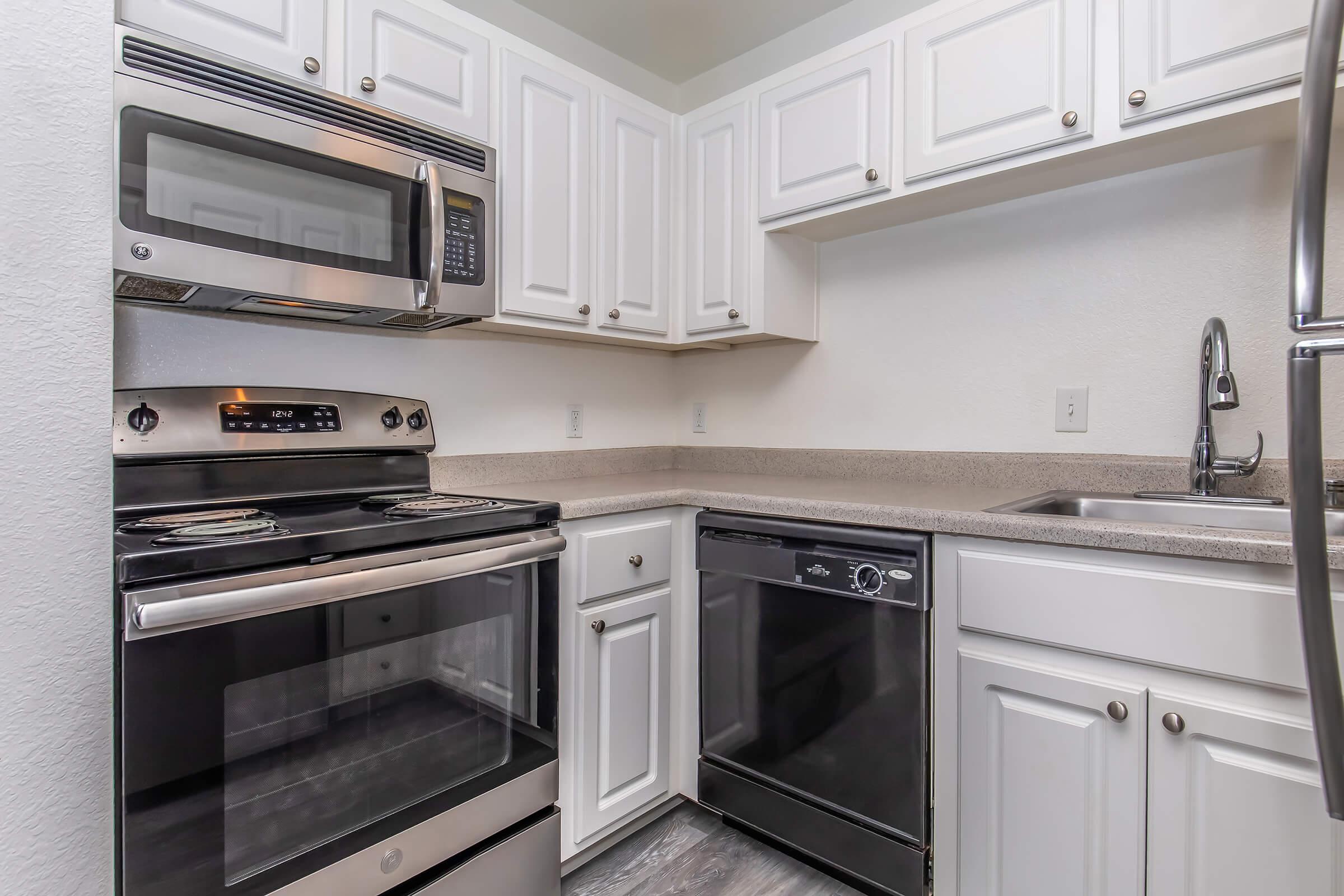 A modern kitchen featuring white cabinetry, stainless steel appliances including a microwave, stove, and dishwasher, with a granite countertop and a sink. The kitchen layout is compact and functional, ideal for small spaces.