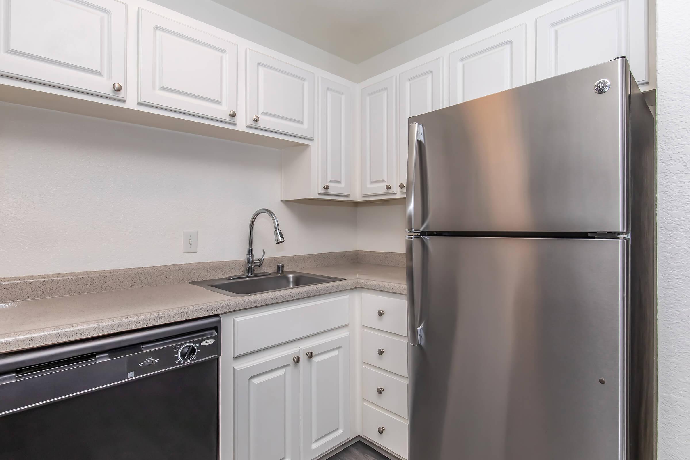 A modern kitchen featuring white cabinets, a stainless steel refrigerator, a dishwasher, and a single basin sink with a sleek faucet. The countertops are a light, neutral color, complementing the cabinetry. The overall design is clean and functional, suitable for compact living spaces.