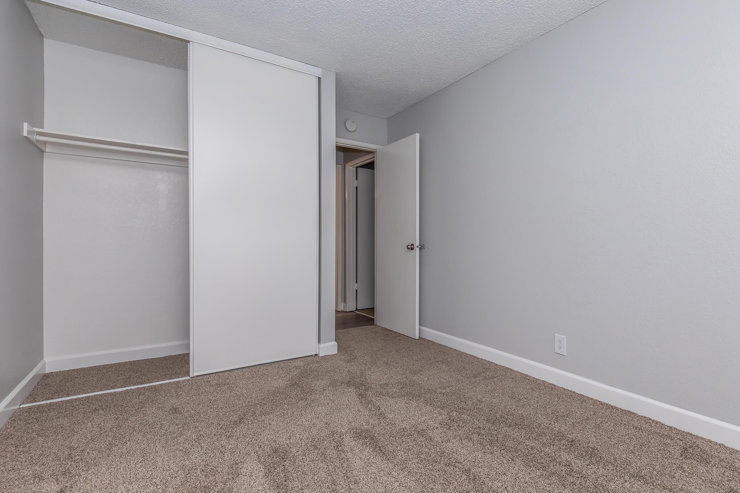 A spacious, empty room with light gray walls and carpeted flooring. There's a large closet with sliding white doors on one side and an open door leading to another room. The overall feel is clean and modern, suitable for various decorating styles.