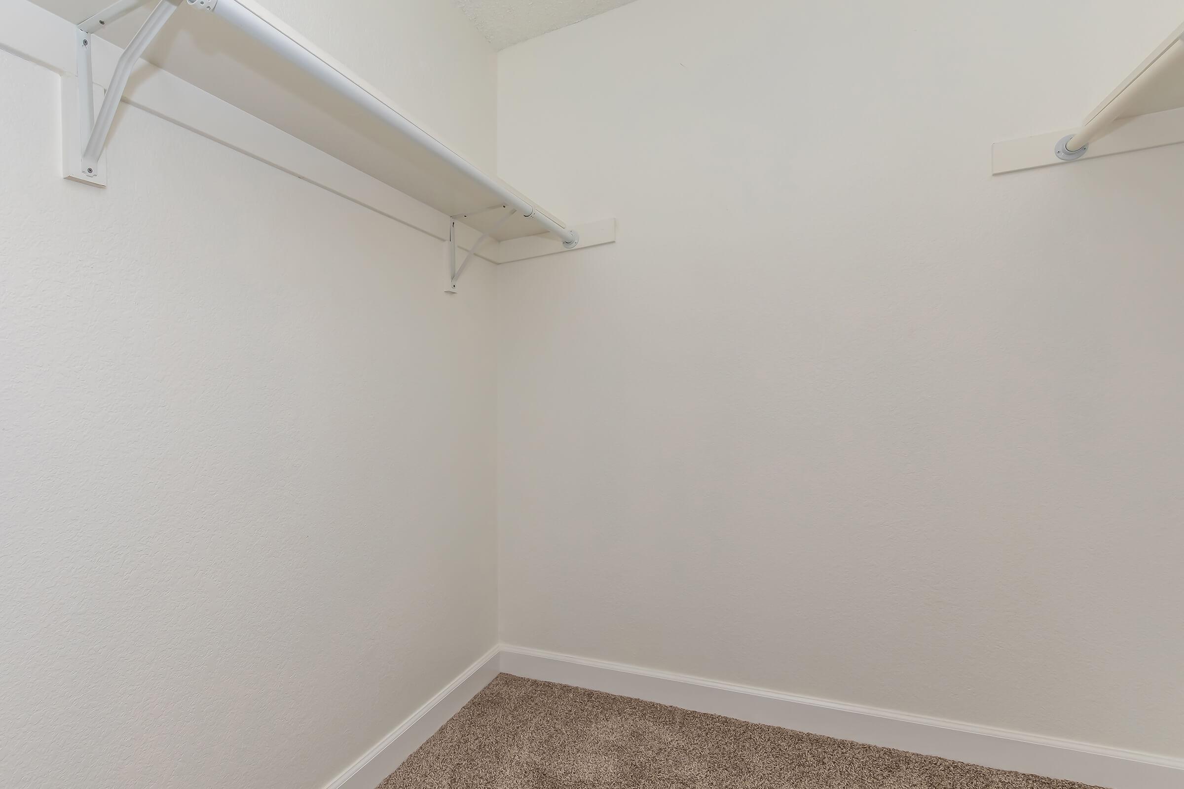Empty closet with two white shelves mounted on the walls. The walls are painted a soft neutral color, and the flooring is covered with a light brown carpet. The space appears well-lit and clean, offering ample storage potential.