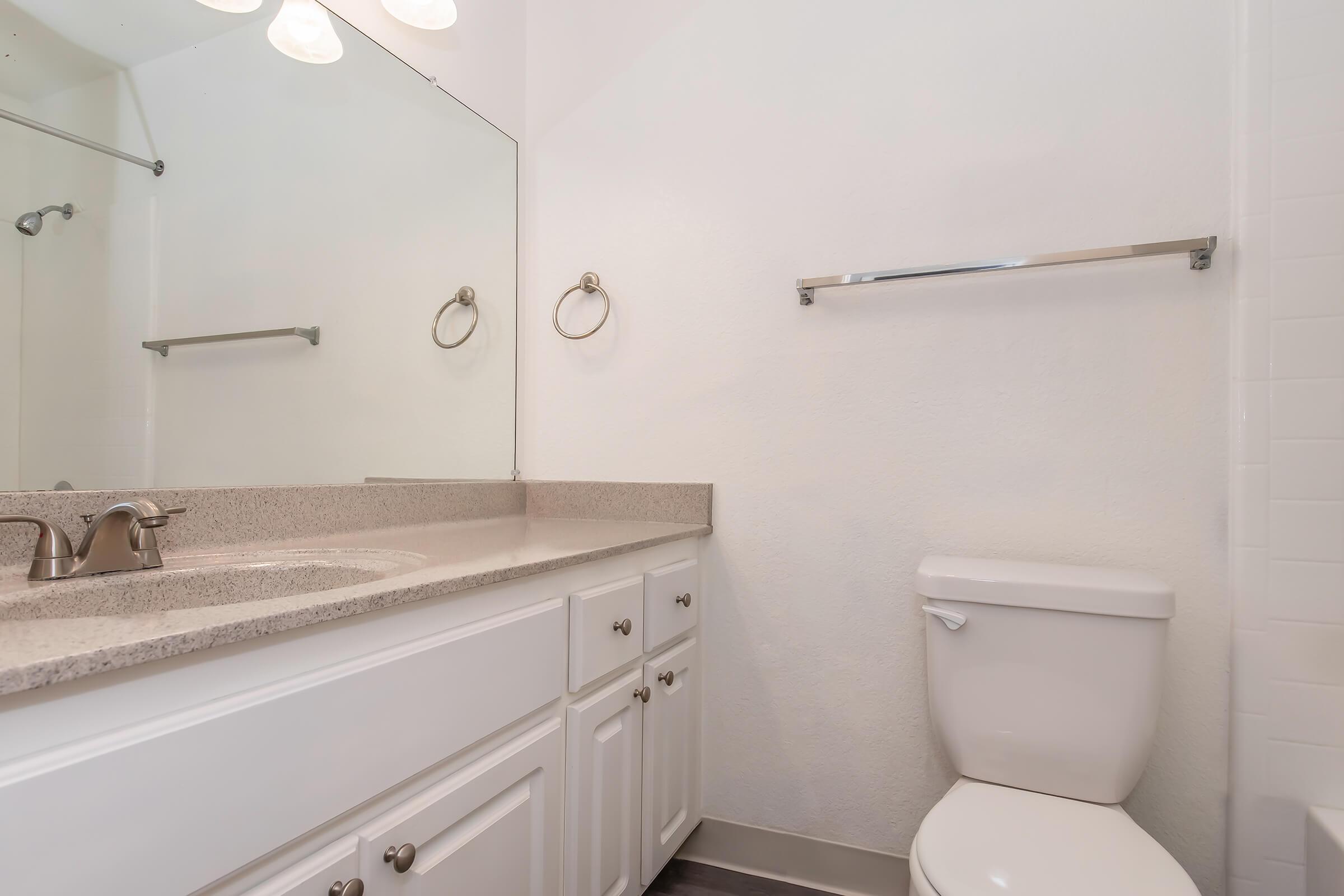 A clean and modern bathroom featuring a white toilet, a counter with a sink, a large mirror, and a towel bar. The walls are painted white, and the flooring is a neutral color, creating a bright and spacious appearance.