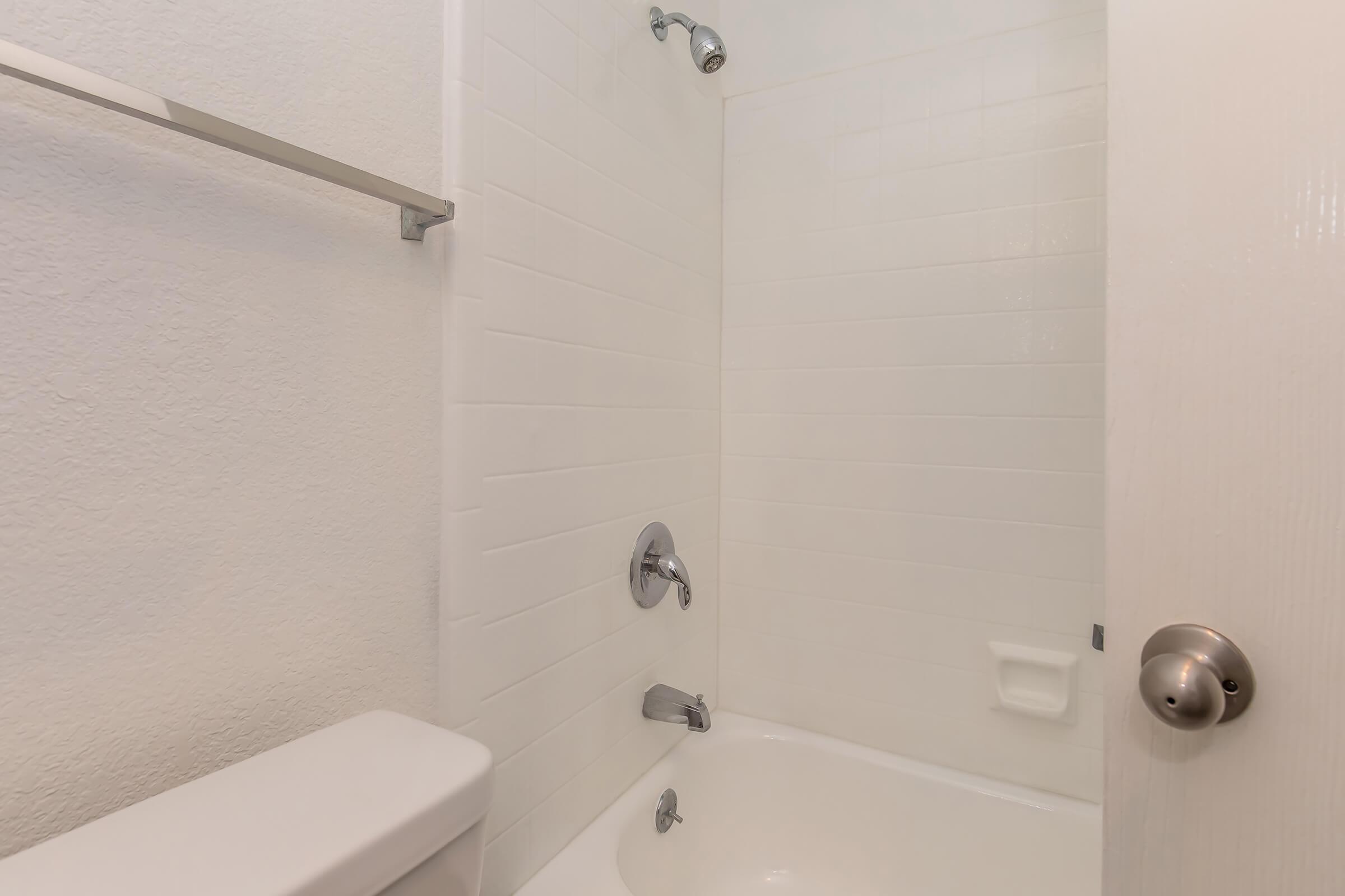 A clean, minimalist bathroom featuring a bathtub with a showerhead, white tiled walls, and a light-colored door. The space is bright and uncluttered, emphasizing simplicity and functionality.