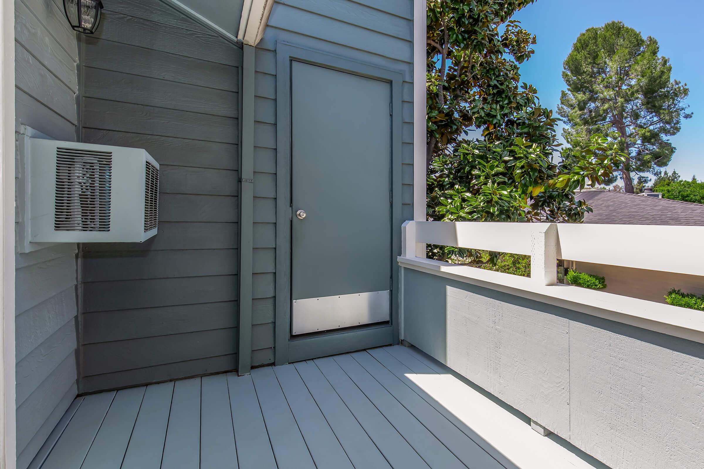 A balcony featuring a gray door, an air conditioning unit mounted on the wall, and wooden railing. The floor is painted white, and there are trees visible in the background under clear blue skies.