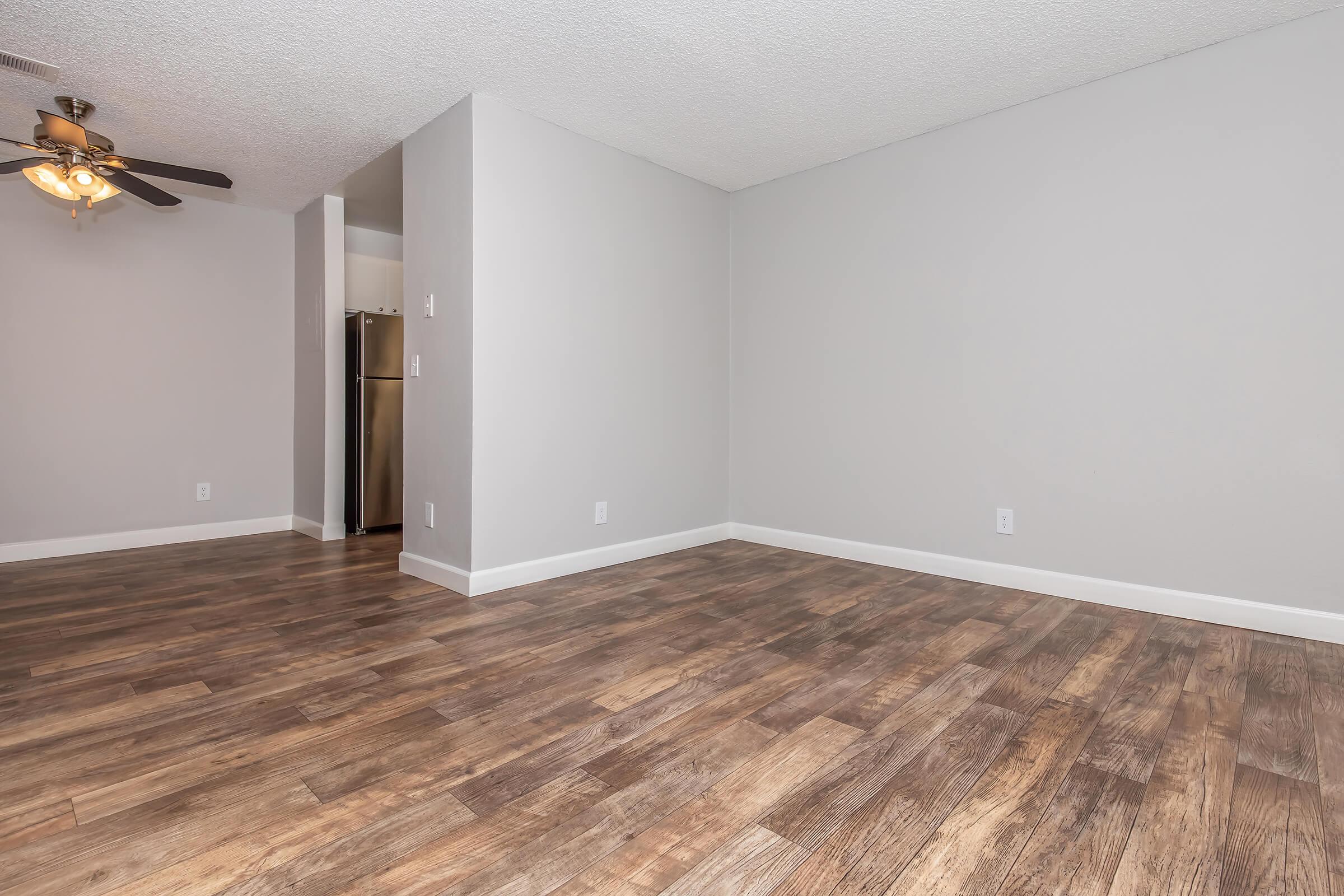 A spacious interior room featuring light-colored walls and a textured ceiling. The floor is covered with wood-like laminate, and there is a ceiling fan with light fixtures. A doorway leads to a kitchen area with a stainless steel refrigerator visible in the background, creating an open and airy atmosphere.