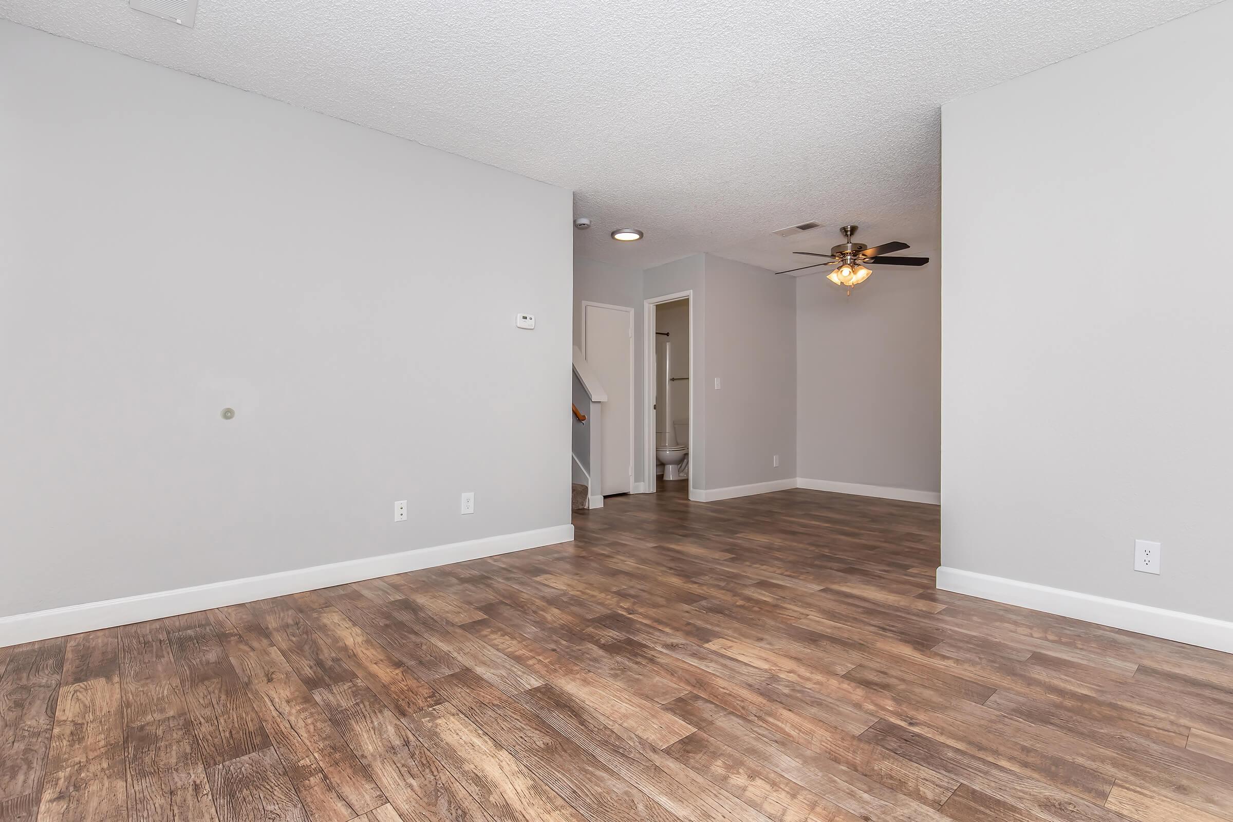 A spacious, modern living area with light gray walls and wood-style flooring. A ceiling fan is visible, and there are doors leading to other rooms, including a bathroom. The layout is open and well-lit, creating a welcoming atmosphere.