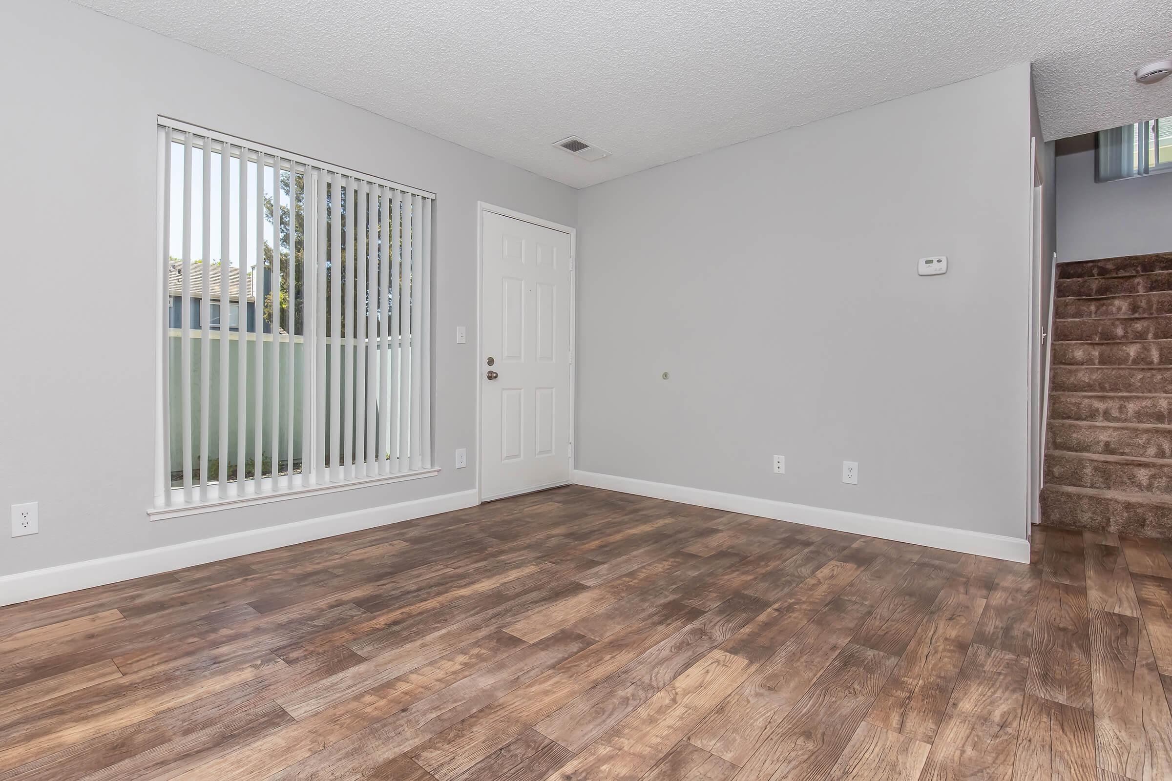 Empty room with wood-like flooring, a white door leading outside, and vertical blinds on a window. The walls are painted light gray, and there is a staircase visible on the right side. The space is bright and minimalist, with no furniture or decorations.