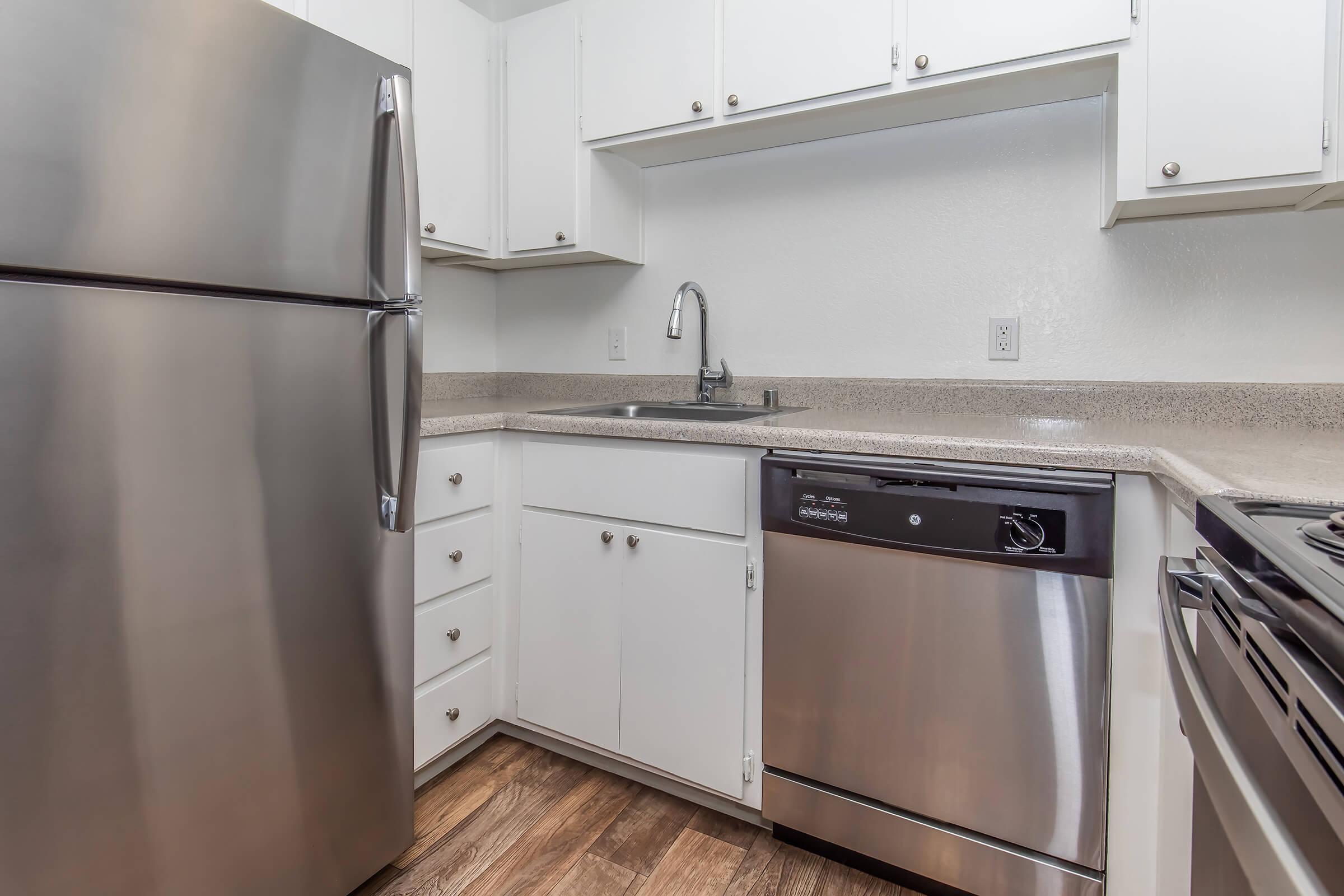 Modern kitchen featuring stainless steel appliances, including a refrigerator and dishwasher. The kitchen has white cabinets, a countertop, a sink with a faucet, and a stove. The flooring is a wood-like laminate, giving the space a clean and contemporary look.