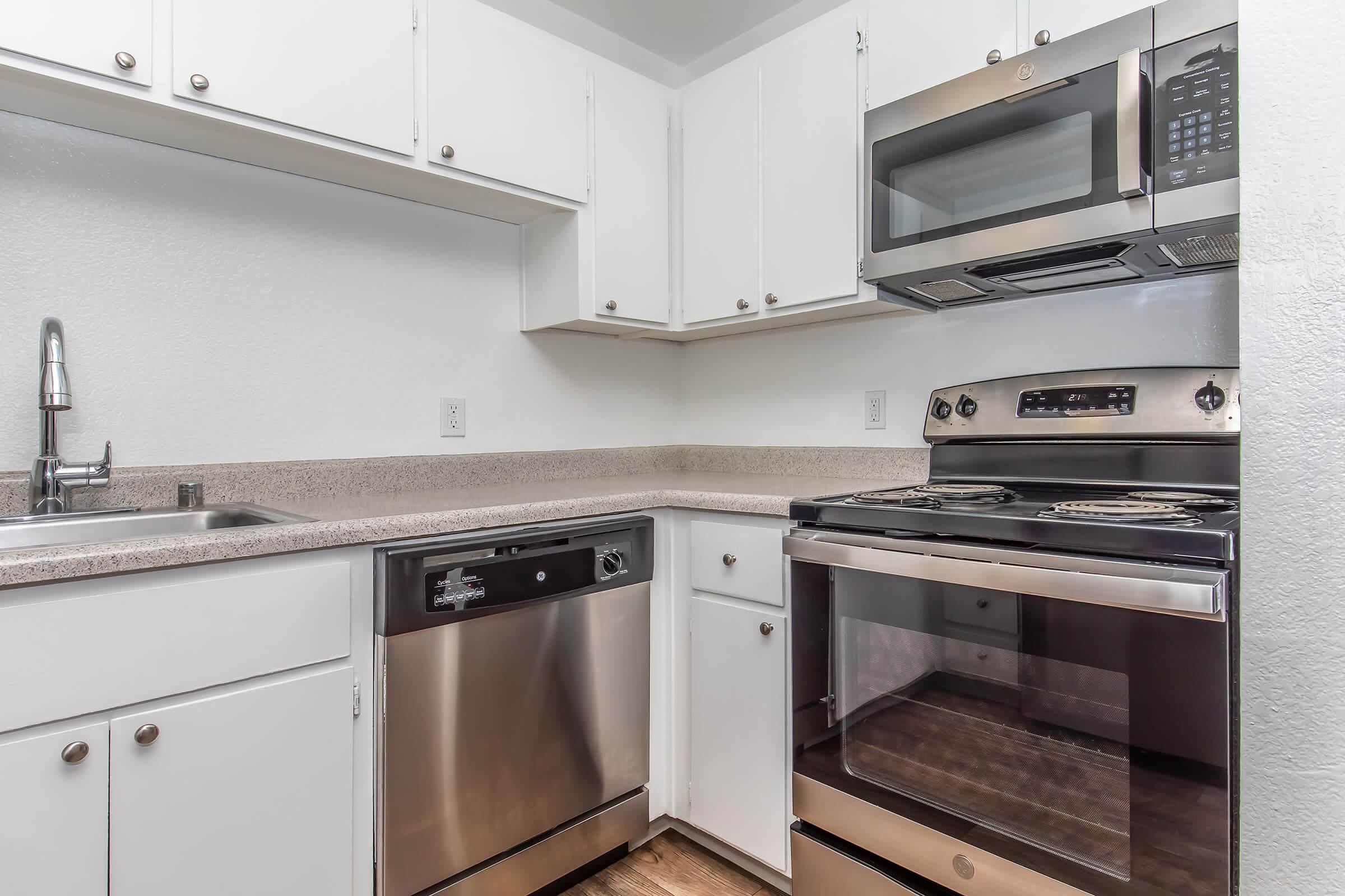 A modern kitchen featuring white cabinets, stainless steel appliances including a dishwasher, microwave, and stove. The countertop is light-colored, and there's a sink with a faucet. The overall design is clean and contemporary, with wood-style flooring.
