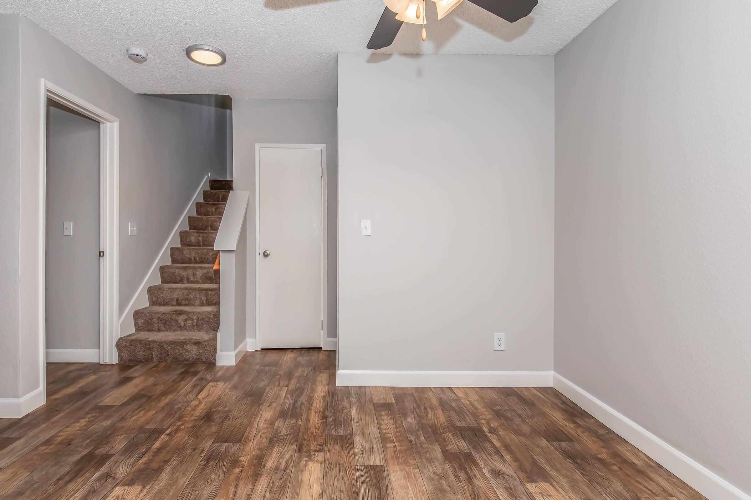 A well-lit, empty living room featuring a ceiling fan, wood-like flooring, and gray walls. A staircase leading to the upper floor is visible on the left, with a closed door at the top. The room has a spacious feel, ideal for arranging furniture and decor.