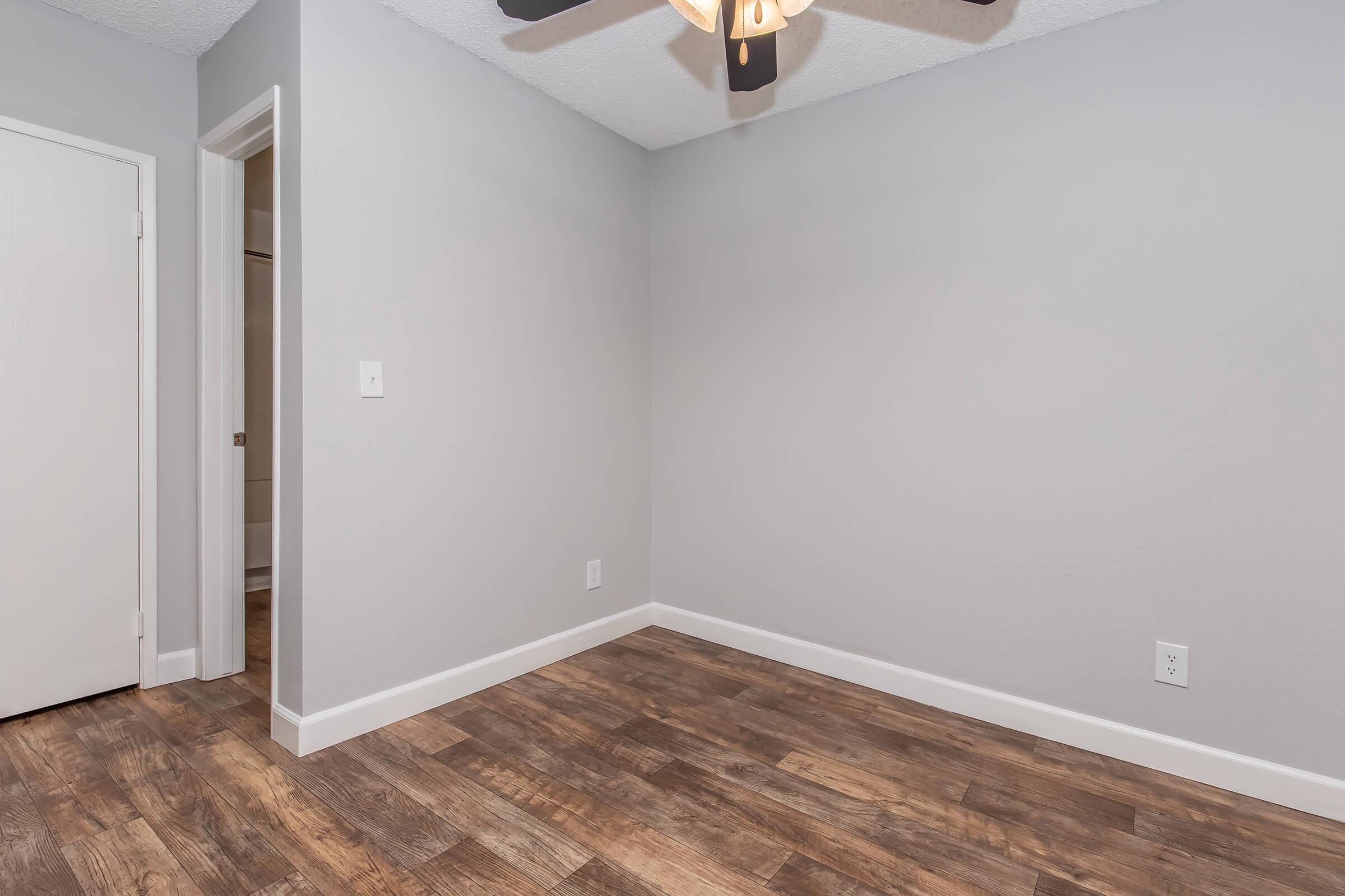 A small, empty room with gray walls and a ceiling fan. The floor is made of brown wooden planks. There are two open doorways: one leading to a bathroom and the other to an adjoining space. The lighting is soft, giving the room a clean and modern appearance.