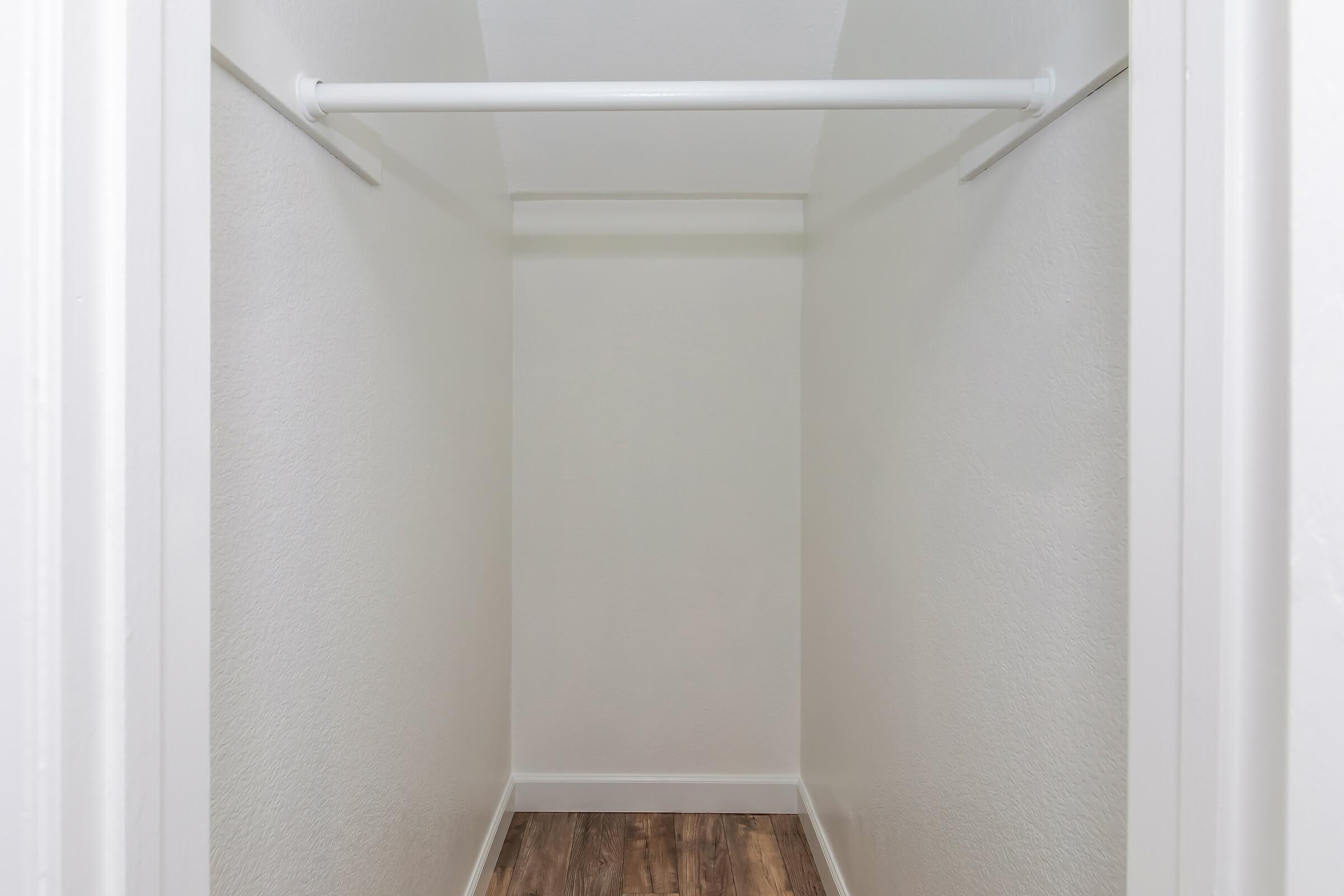 A vacant closet with white walls, a white hanging rod, and a wooden floor. The space is well-lit and empty, providing ample room for storage or organization.