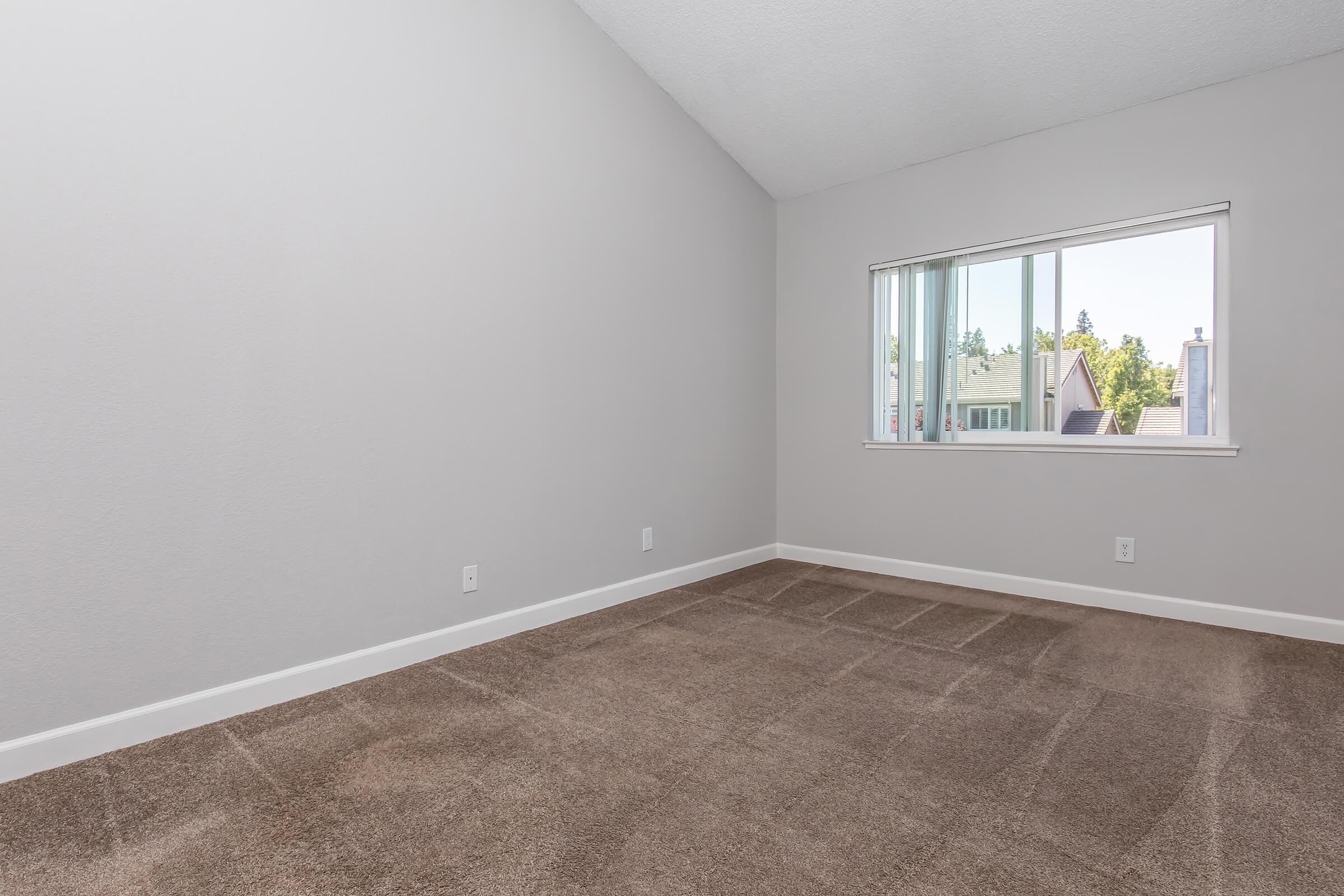 A minimalistic room with light gray walls and a sloped ceiling. The floor is covered in soft brown carpet. There is a large window on one wall, allowing natural light to enter and providing a view of rooftops outside. The space is uncluttered and ready for furnishings.