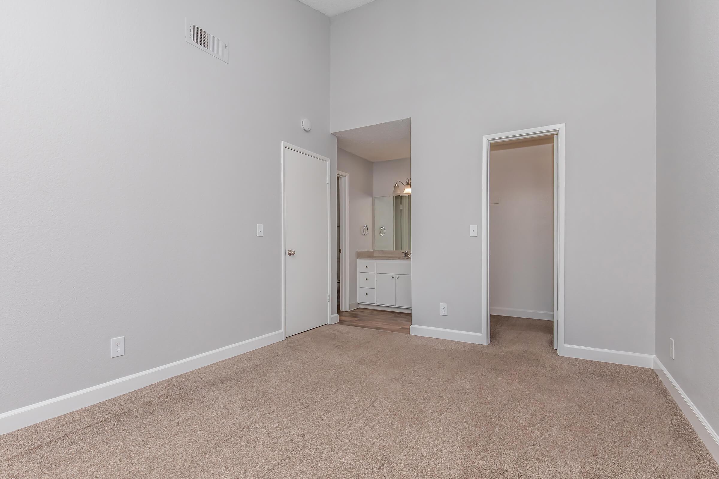 A spacious, empty room with light gray walls, carpeted flooring, and two doorways leading to other areas. One doorway opens to a bathroom with a vanity and mirror visible. Natural light brightens the space, creating a clean and modern atmosphere.