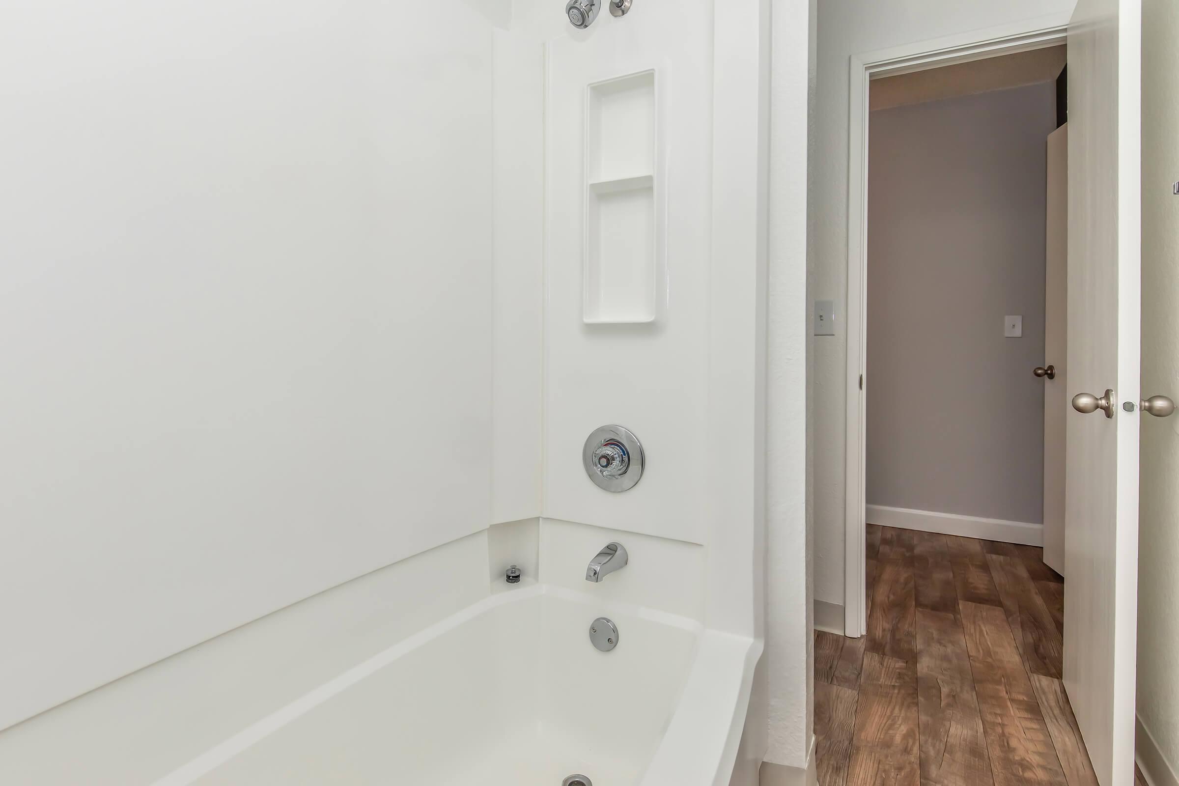 A modern bathroom featuring a white bathtub with a sleek design, a wall-mounted shower fixture, and built-in shelving. The room has neutral-colored walls and a wooden floor, with a doorway visible in the background leading to another room.