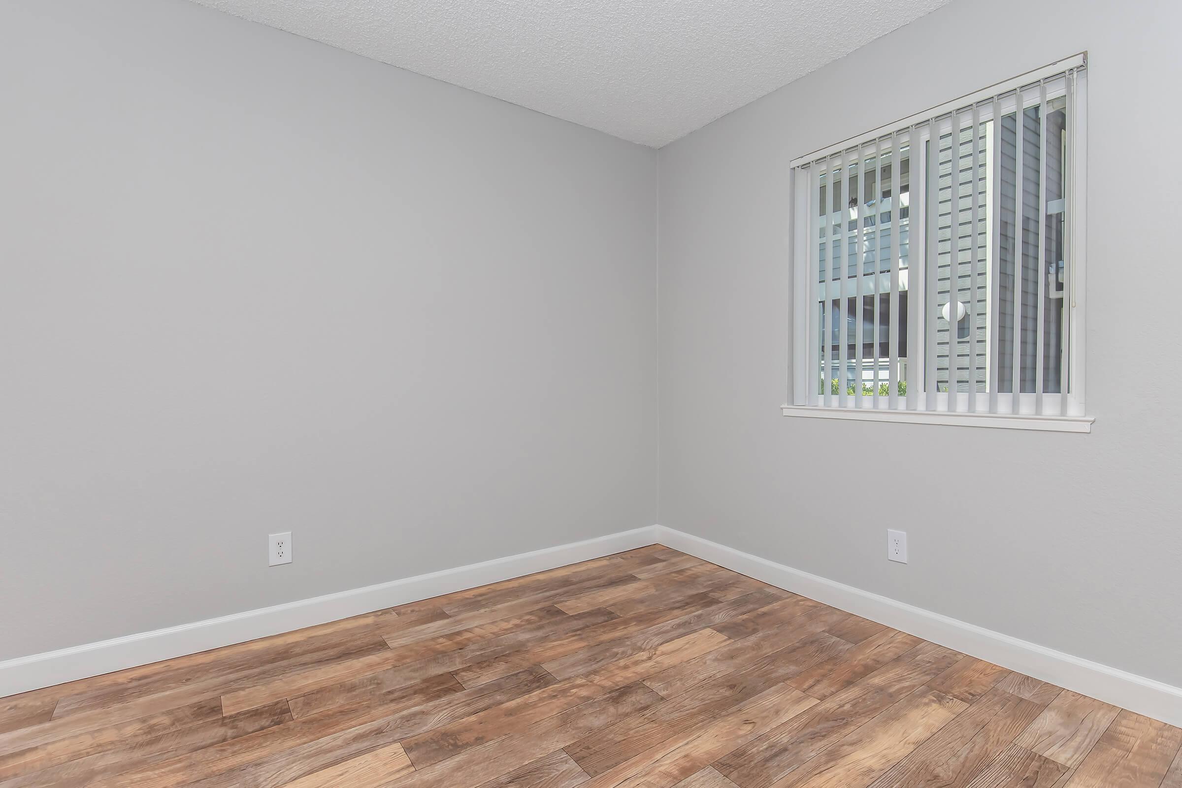 Empty interior of a room with light gray walls and wooden flooring. A small window with white blinds lets in natural light. The space is unfurnished and minimalistic, creating an open and inviting atmosphere.