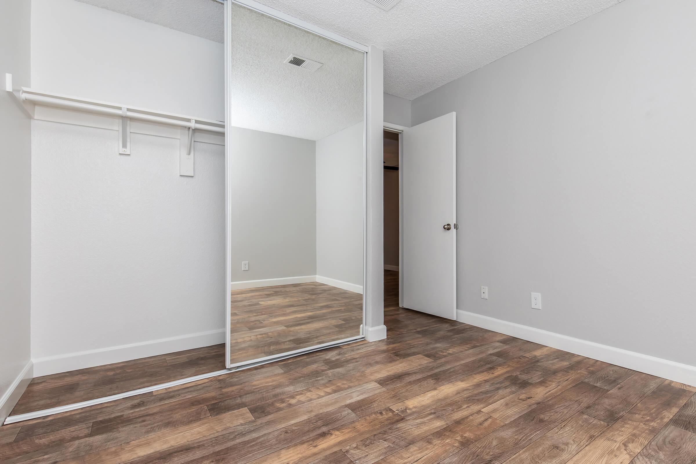 Empty room with gray walls and a mirrored closet door. Light wood-like flooring. A single door leads to another area, and there are no furnishings or decorations. The space is bright and uncluttered, suggesting potential for various uses or personal decoration.