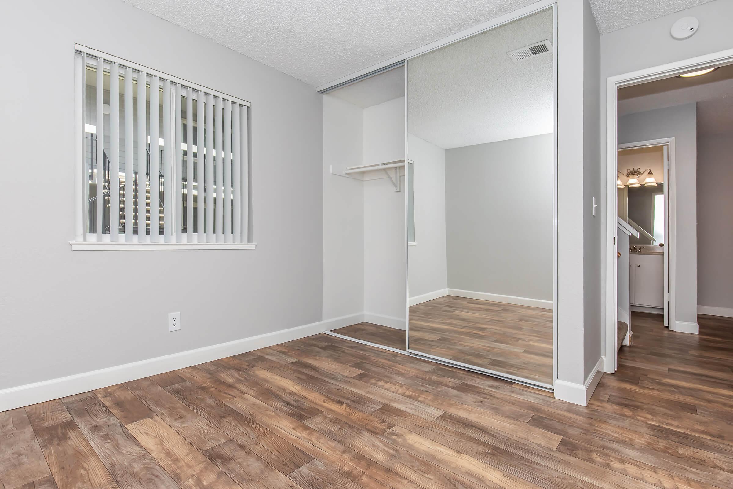 Empty room with wooden flooring, light gray walls, and a large mirror closet. A window with vertical blinds lets in natural light. A doorway leads to another room, possibly a bathroom, indicated by soft lighting in the background. The space appears clean and modern, suitable for various interior designs.