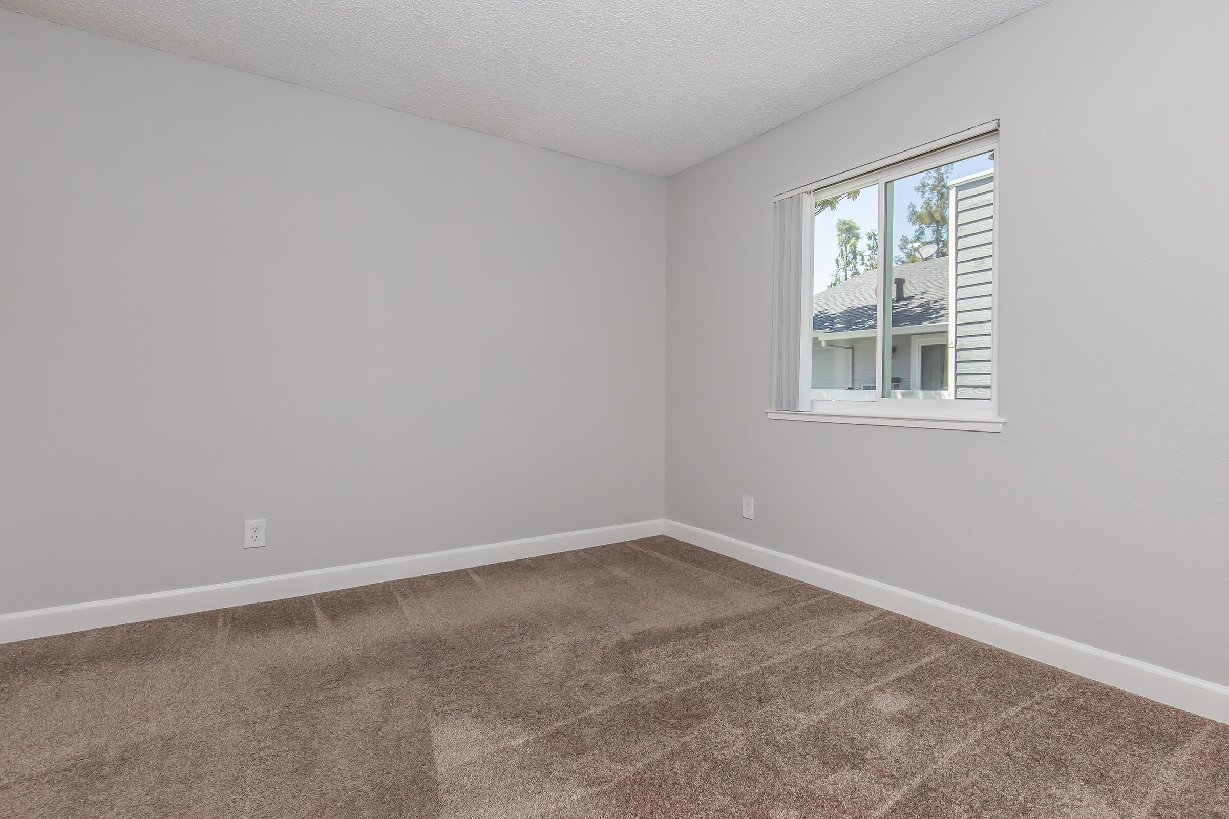Empty room with light gray walls and a window. The floor is covered in beige carpet. There is no furniture, creating a spacious and minimalist feel. Natural light enters through the window, illuminating the area.