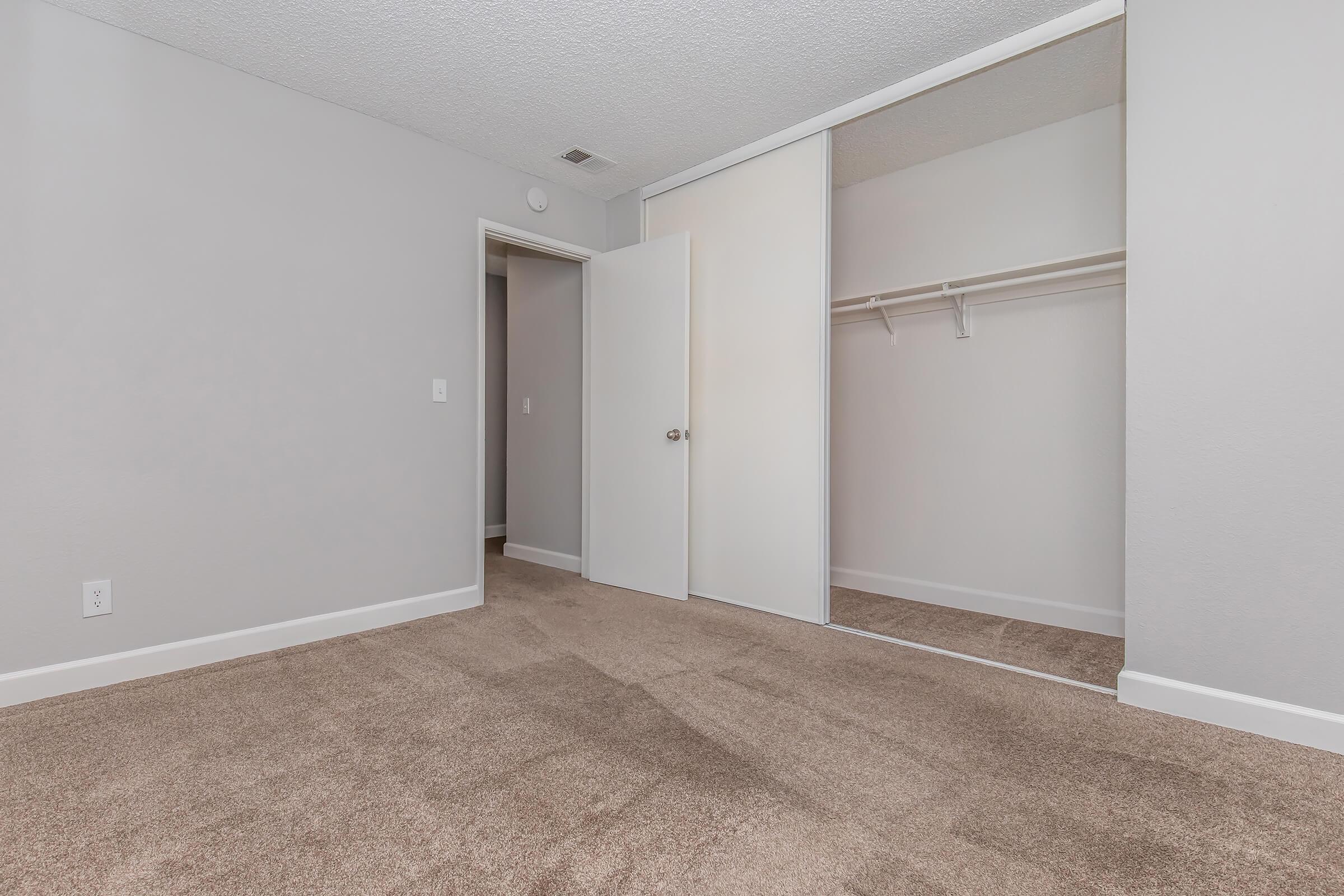 A spacious, empty room with light gray walls and carpet. It features a mirrored closet with sliding doors, a doorway leading to another room, and a white door. The overall ambiance is bright and clean, perfect for personalization.