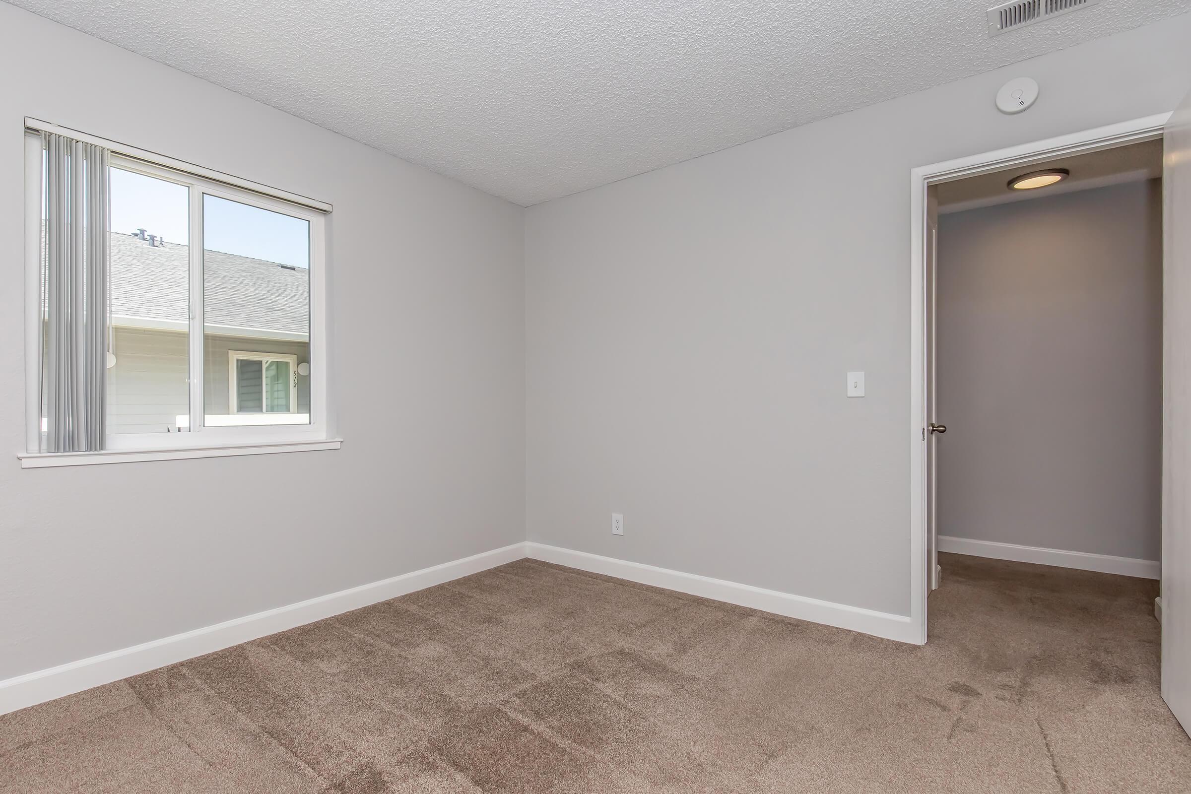 A vacant room with beige carpet and light gray walls. There's a large window with vertical blinds letting in natural light, and a door leading to a hallway. The space is empty and well-lit, suitable for various uses.