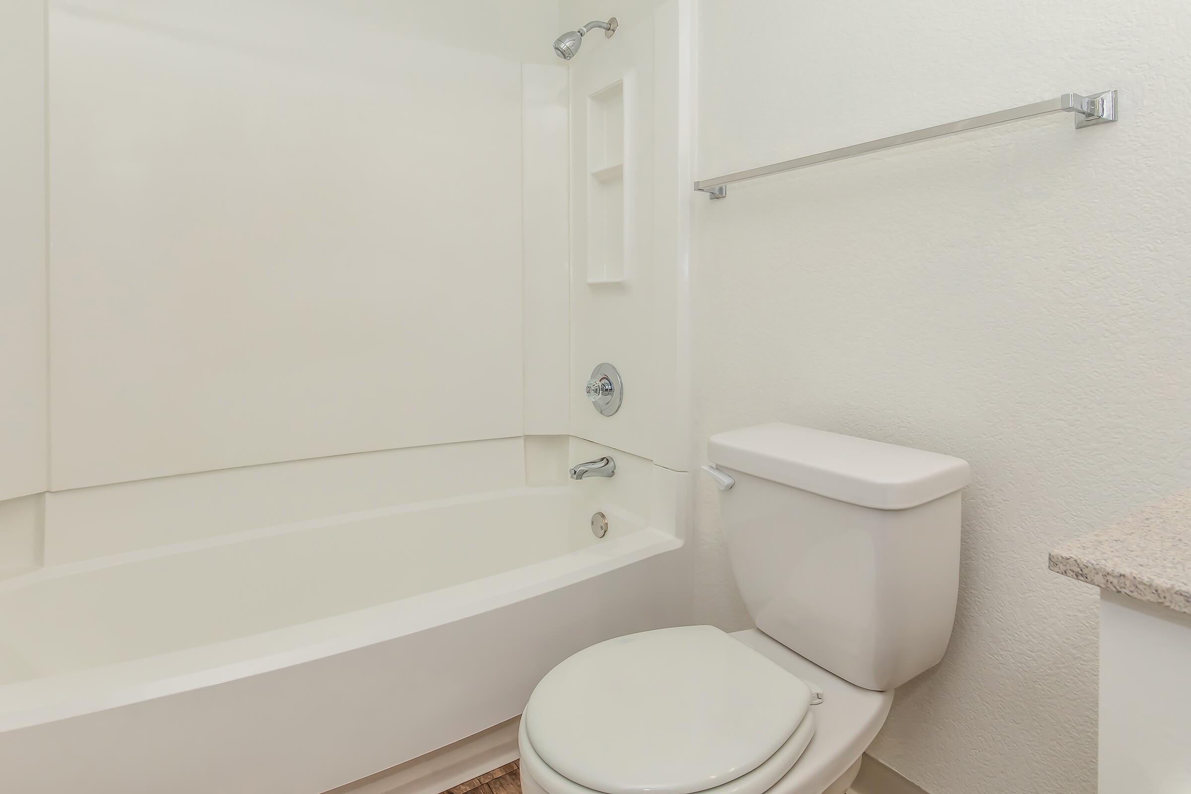 A clean, bright bathroom featuring a white bathtub with a shower head, a wall-mounted towel rack, and a white toilet. The walls are painted in a light color, and there is a countertop visible, suggesting a tidy and modern space.