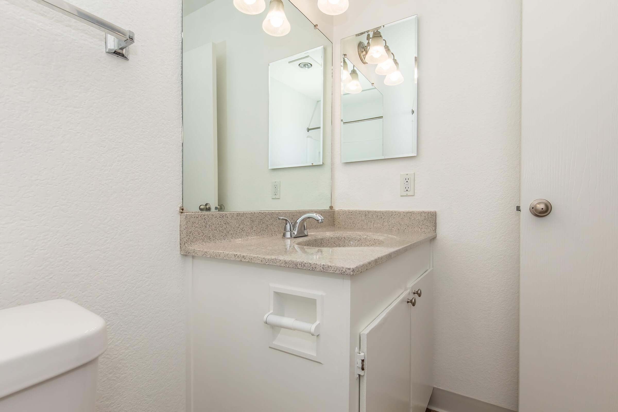 A clean and modern bathroom featuring a white sink and countertop, large mirrors, and bright overhead lighting. The walls are painted in a light color, and there is a toilet in the corner. The overall aesthetic is minimalist and well-lit, creating a spacious feel.