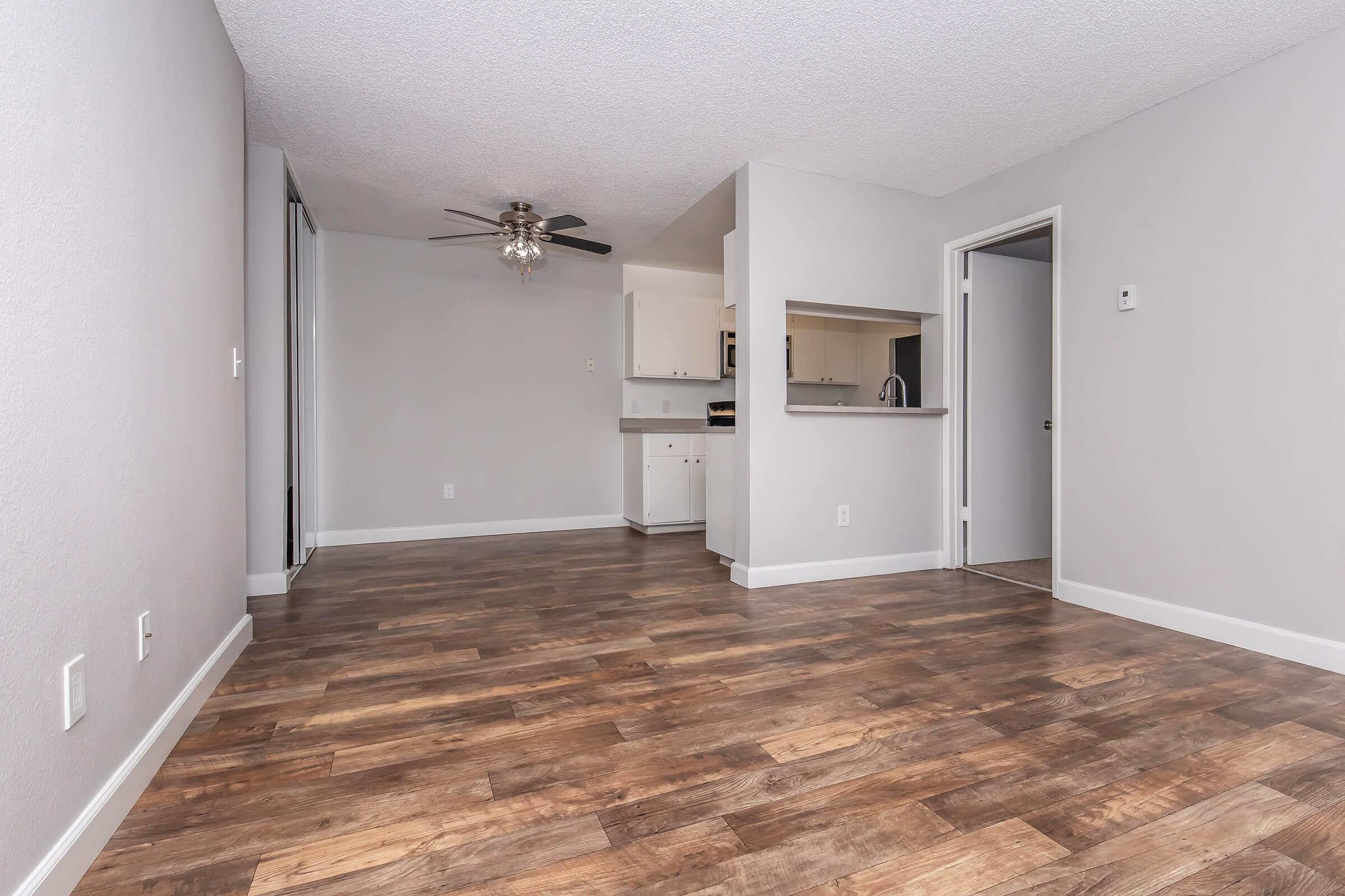 A spacious, modern apartment interior featuring a wooden floor, light gray walls, and a ceiling fan. There is an open layout with a doorway leading to a kitchen area in the background. Natural light fills the room, highlighting the clean design and contemporary feel.