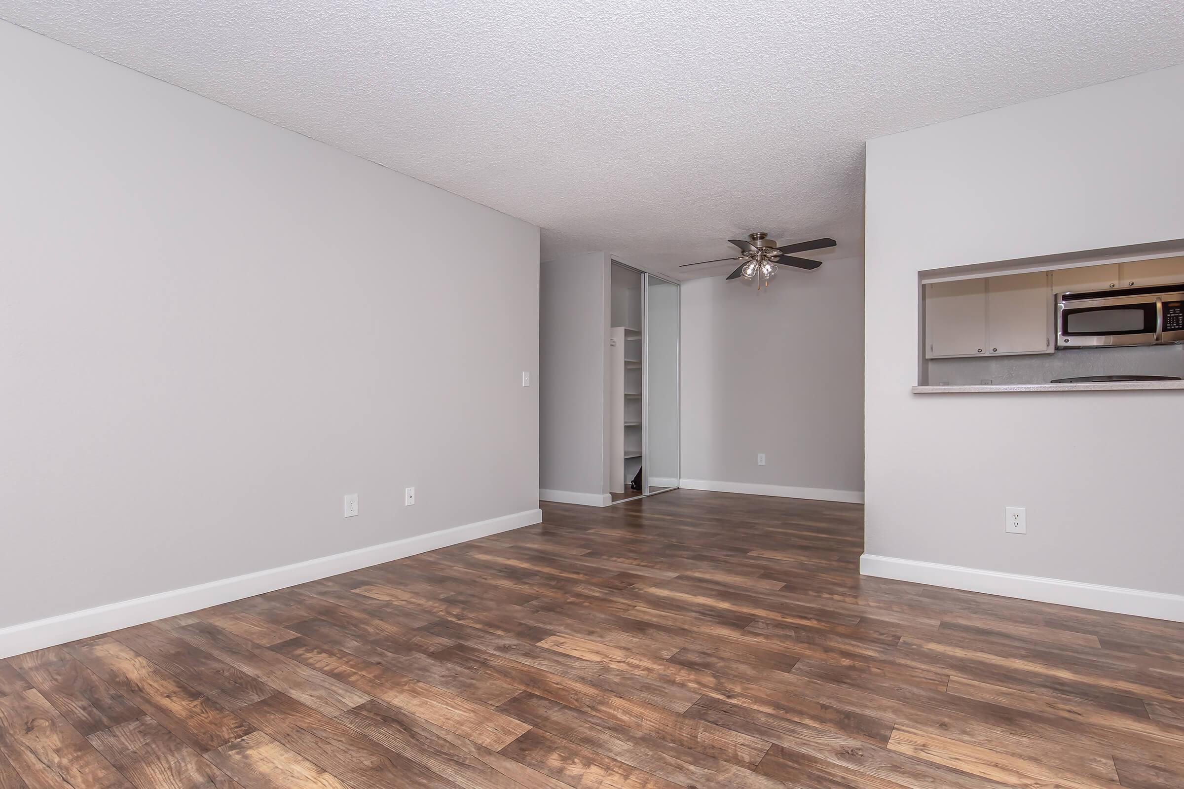 Empty apartment interior featuring light gray walls, a ceiling fan, and laminate wood flooring. An open layout leads to a small kitchen area with a microwave visible. There is a closet space in the background, providing additional storage. The room has a bright and spacious feel.