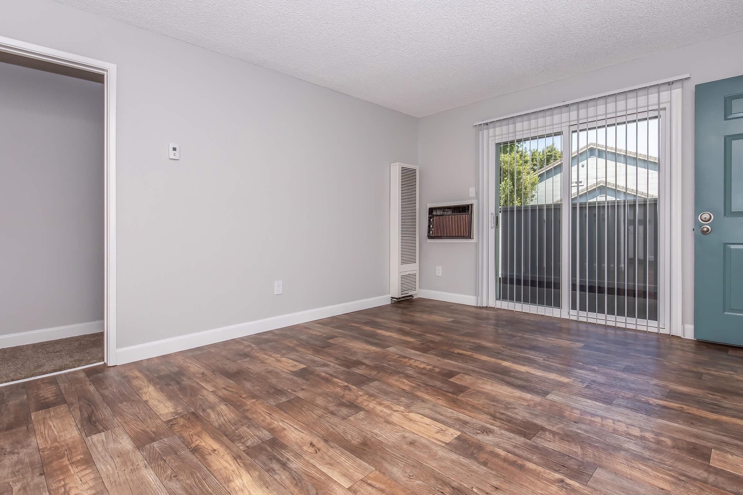 A spacious, empty room with light gray walls and a wood-like floor. There's a sliding glass door leading to an outdoor area, a large window, and a wall-mounted heating unit. An adjacent room is partially visible, and the interior is well-lit with natural light.