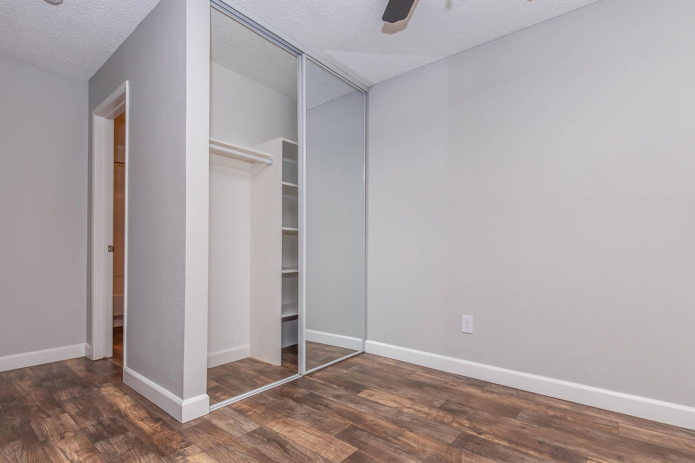 A well-lit room featuring hardwood-style flooring, a mirrored closet door, and a neutral gray wall. The space includes an open doorway leading to another area, suggesting a functional layout suitable for various purposes.