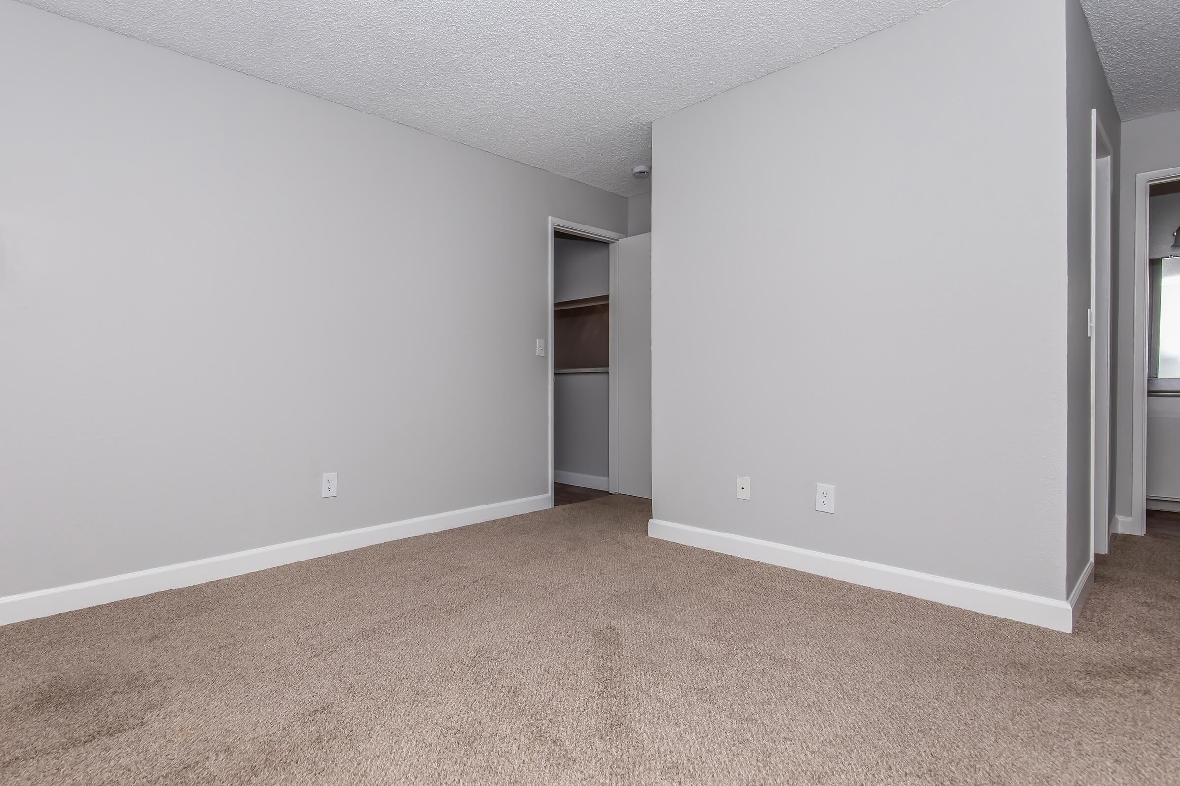 Empty room with beige carpet and light gray walls. A closet is visible on the left side, with a door partially open. The overall ambiance is neutral and spacious, with no furniture present. A doorway to another room can be seen in the background on the right.