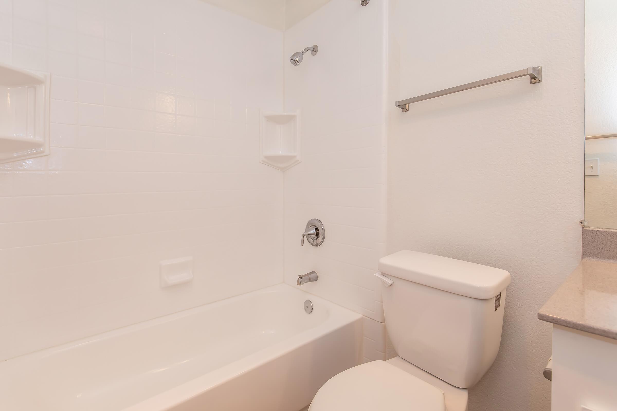 A clean, modern bathroom featuring a white bathtub with a showerhead, soap niches on the wall, a white toilet, and a small countertop area. The walls are tiled in a simple white finish, creating a bright and spacious appearance.
