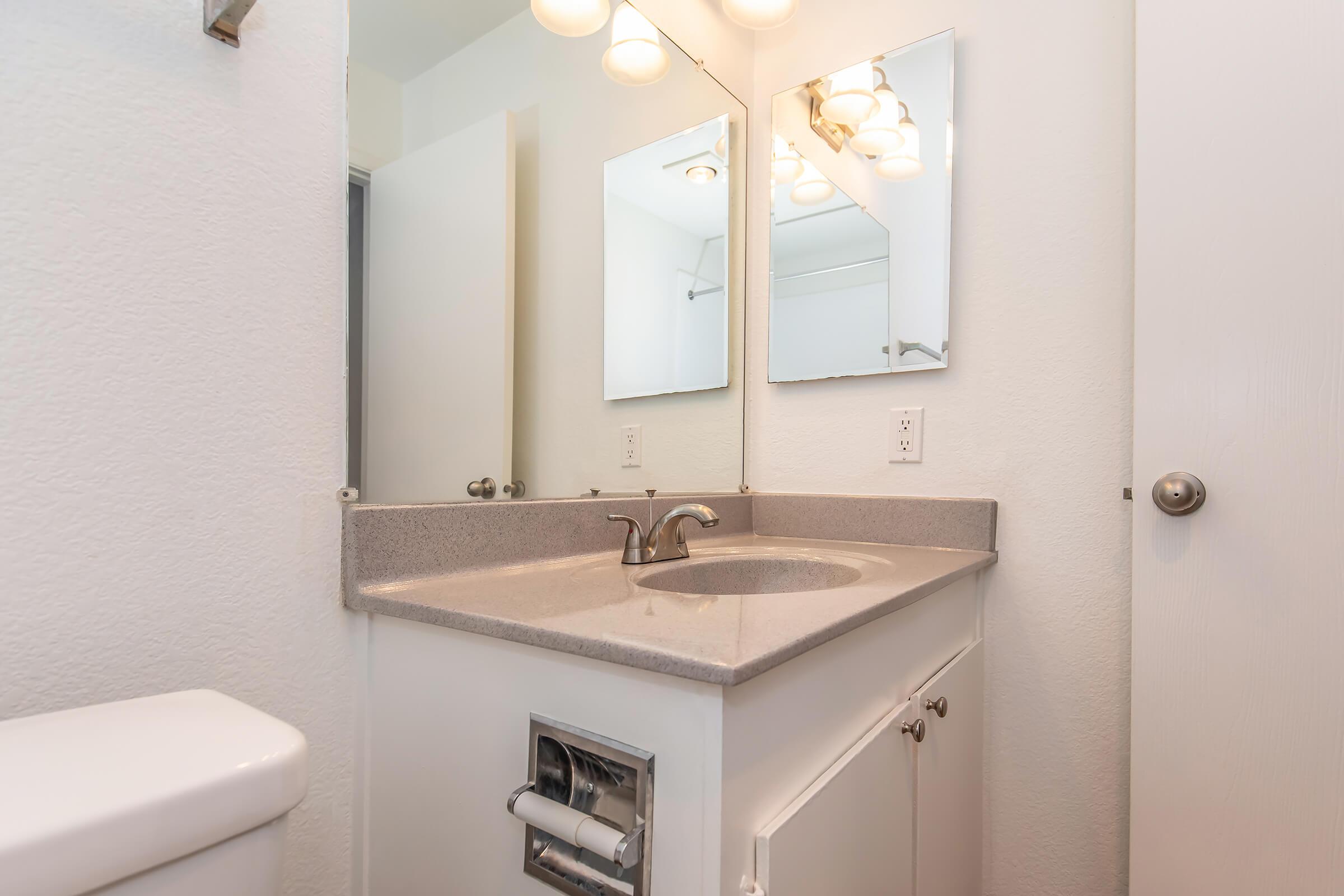 A clean, modern bathroom with a white sink and countertop, two mirrors above the sink, and a toilet to the left. The walls are painted light colors, and there is a bright lighting fixture above the mirrors. A door is partially visible, indicating access to another area.