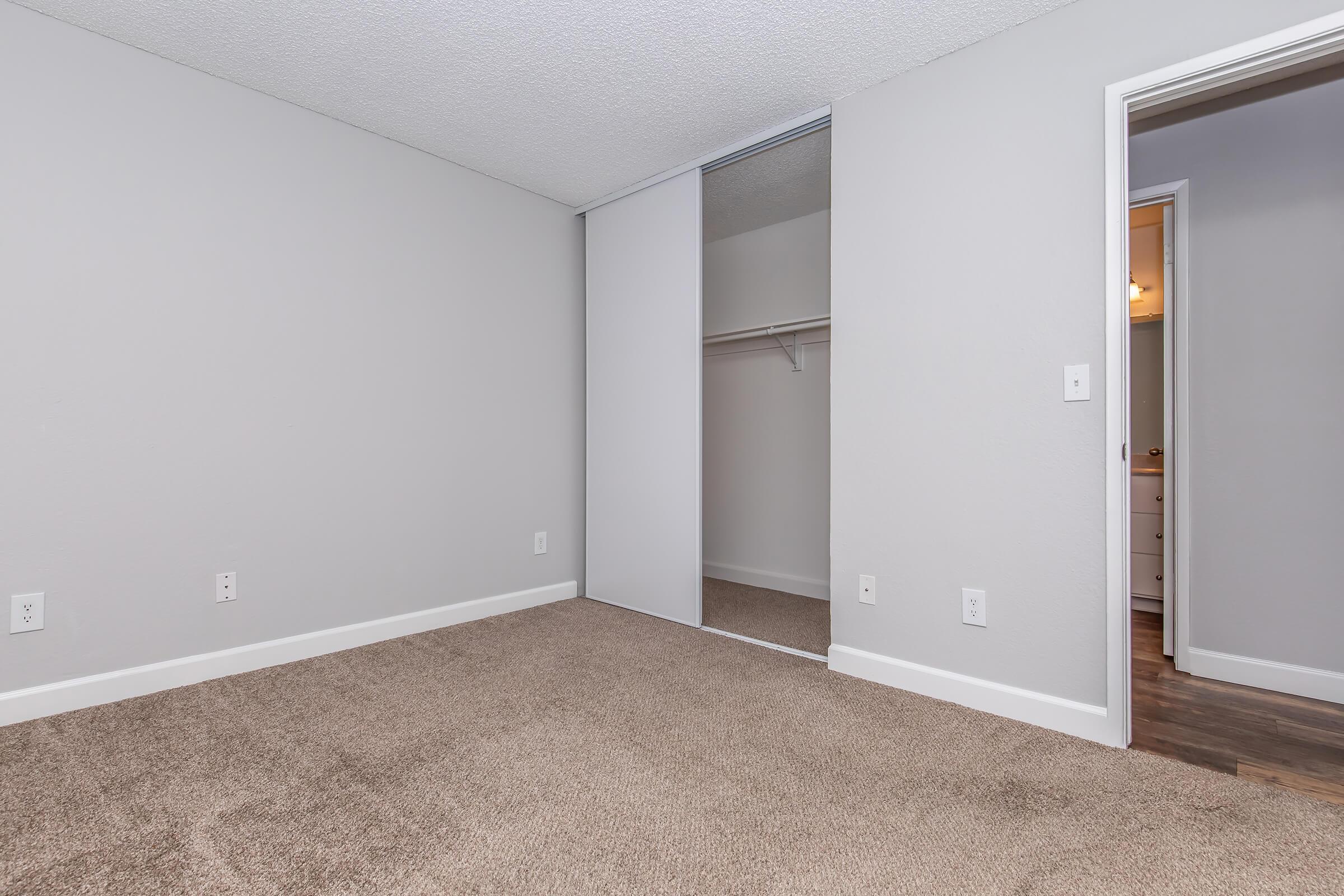 A minimalistic bedroom with light gray walls and a large closet featuring sliding mirrored doors. The floor is covered with carpet, and there is a doorway visible in the background leading to another room. The overall appearance is clean and uncluttered, with neutral colors.