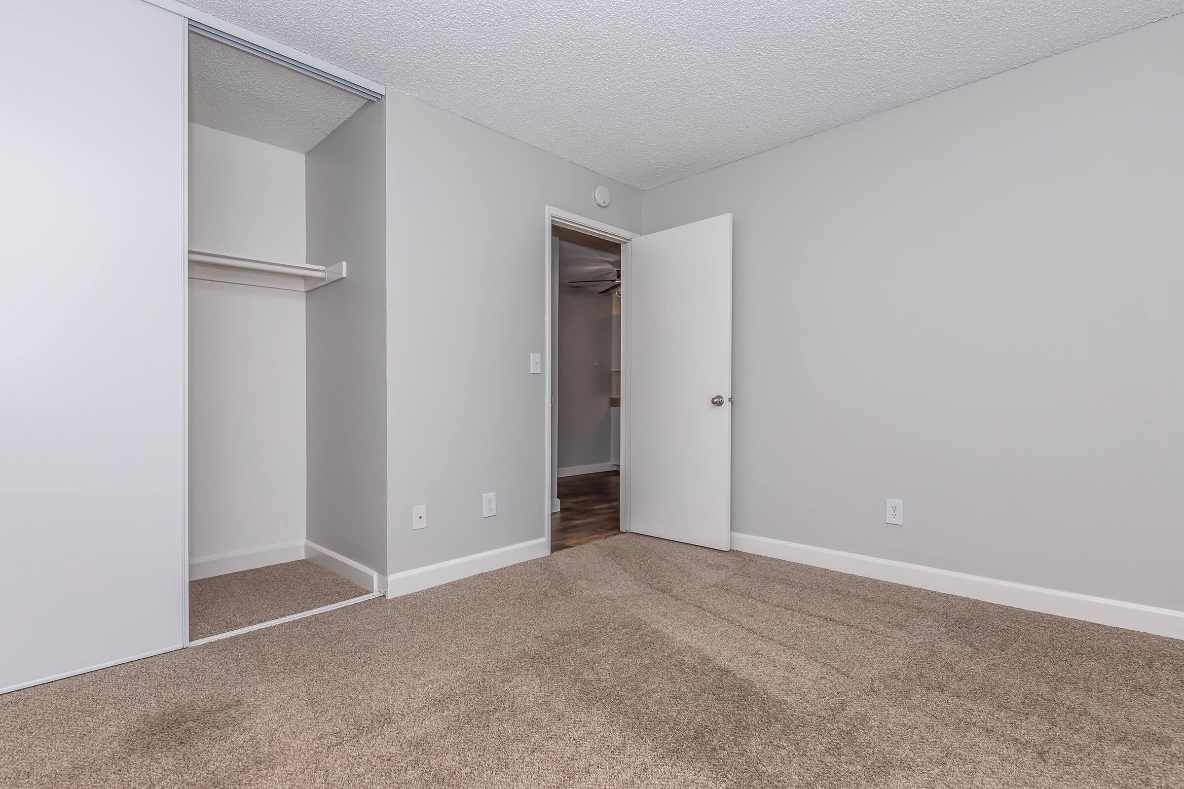 Empty room featuring light gray walls and beige carpet. A closet with a sliding mirror door is visible on the left, and a closed white door leads to another space. The room appears well-lit and spacious, with a simple and modern design.