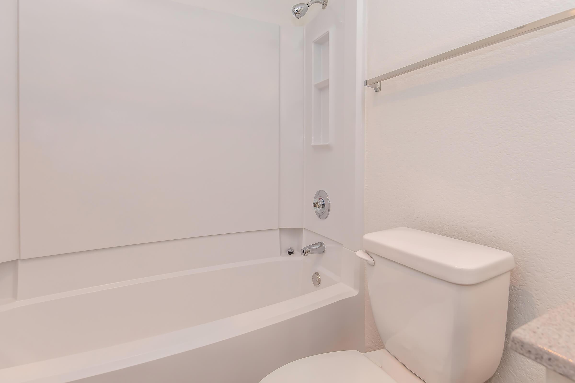 A clean, modern bathroom featuring a white bathtub with a minimalist design, a white toilet, and light-colored walls. The bath area is well-lit, giving a spacious and tidy appearance, with a towel bar mounted on the wall nearby.