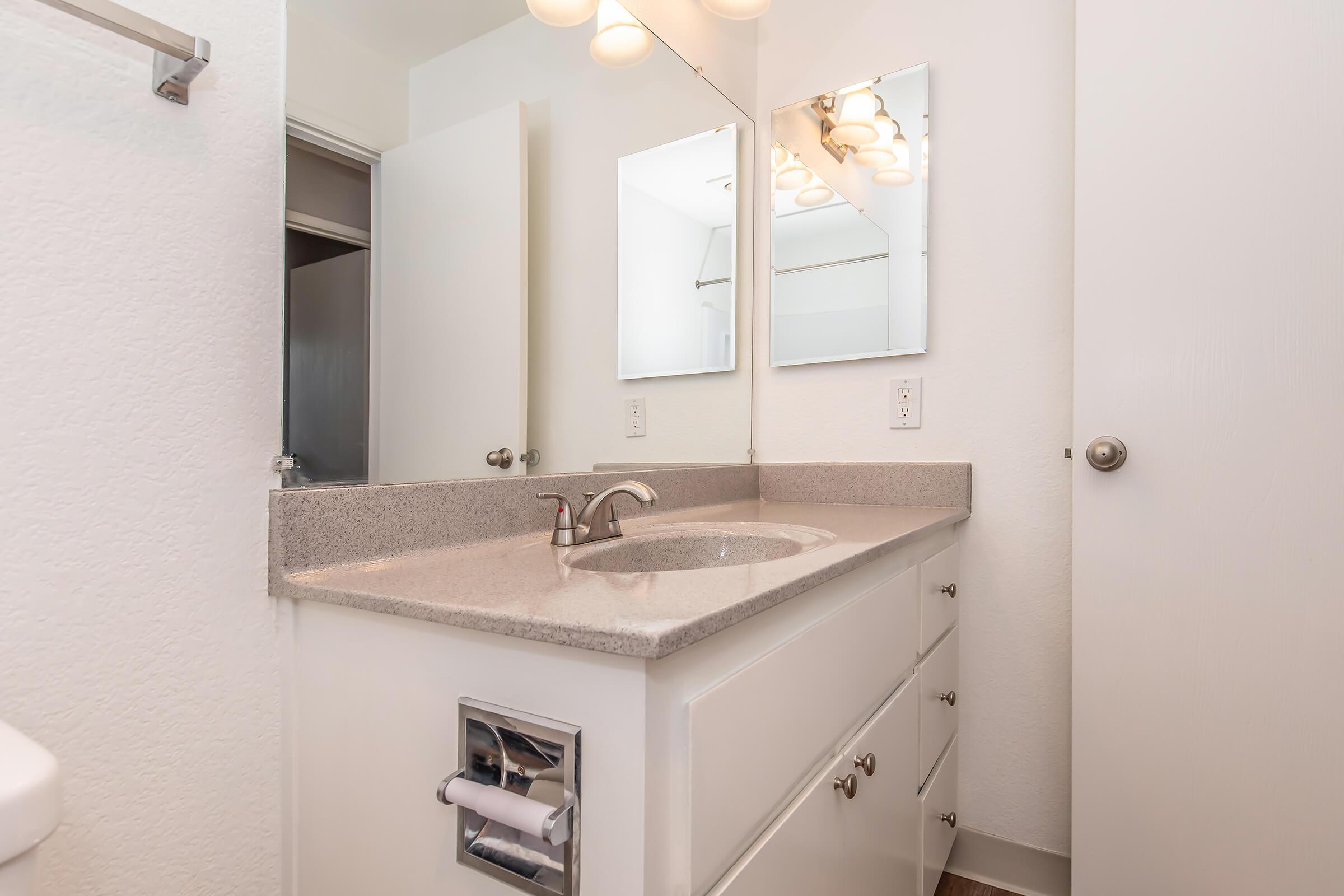 A modern bathroom featuring a light-colored vanity with a sink, two mirrors above it, and a light fixture. The walls are painted white, and there’s a door leading to another room visible in the background. The floor has a wooden finish, and a toilet is partially visible to the left.