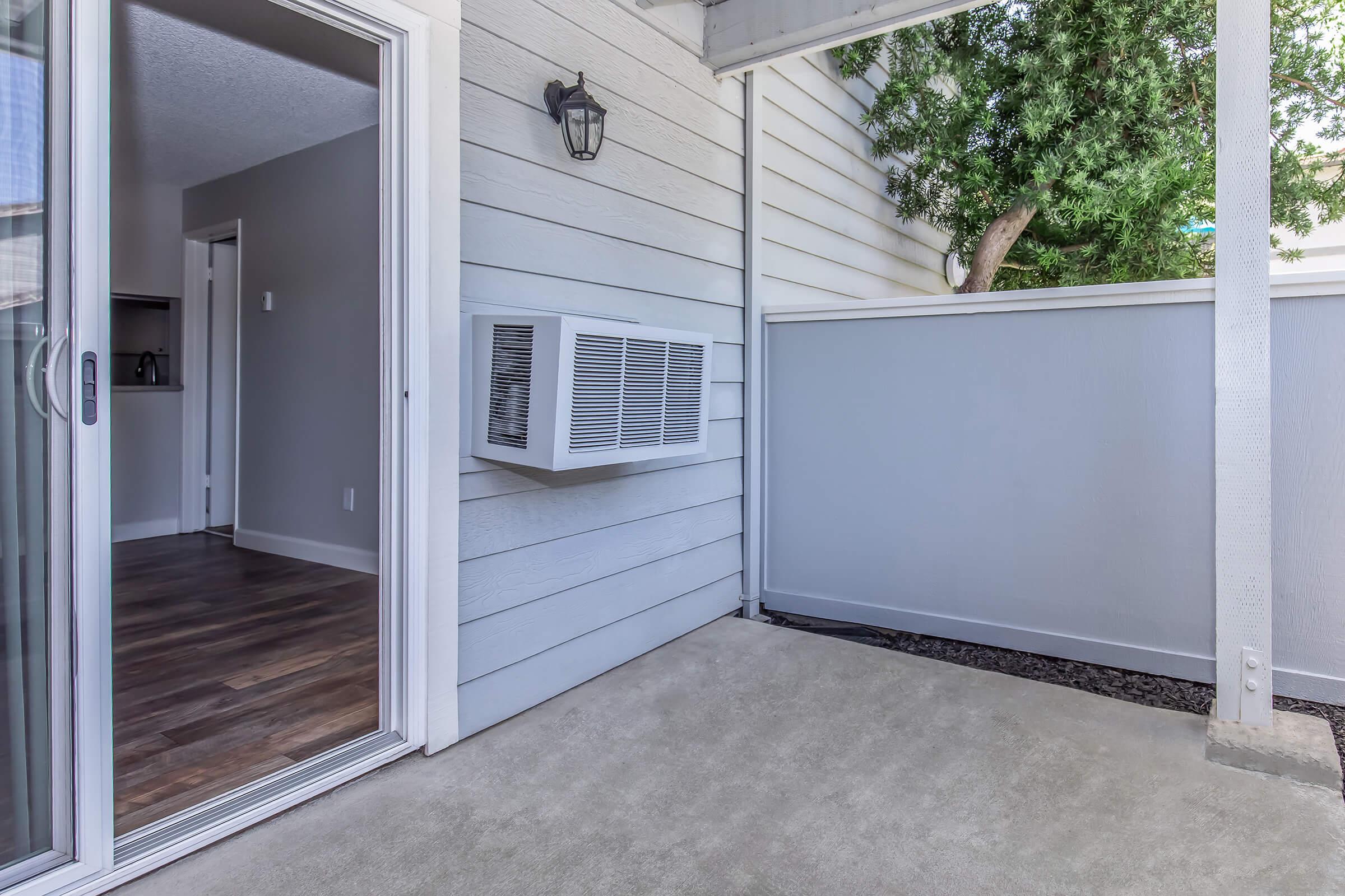 A corner view of a patio area featuring a sliding glass door leading into a room, an air conditioning unit mounted on the wall, and a grey fence. The patio is partially shaded with a tree visible in the background. The flooring is concrete, and the walls are light-colored.
