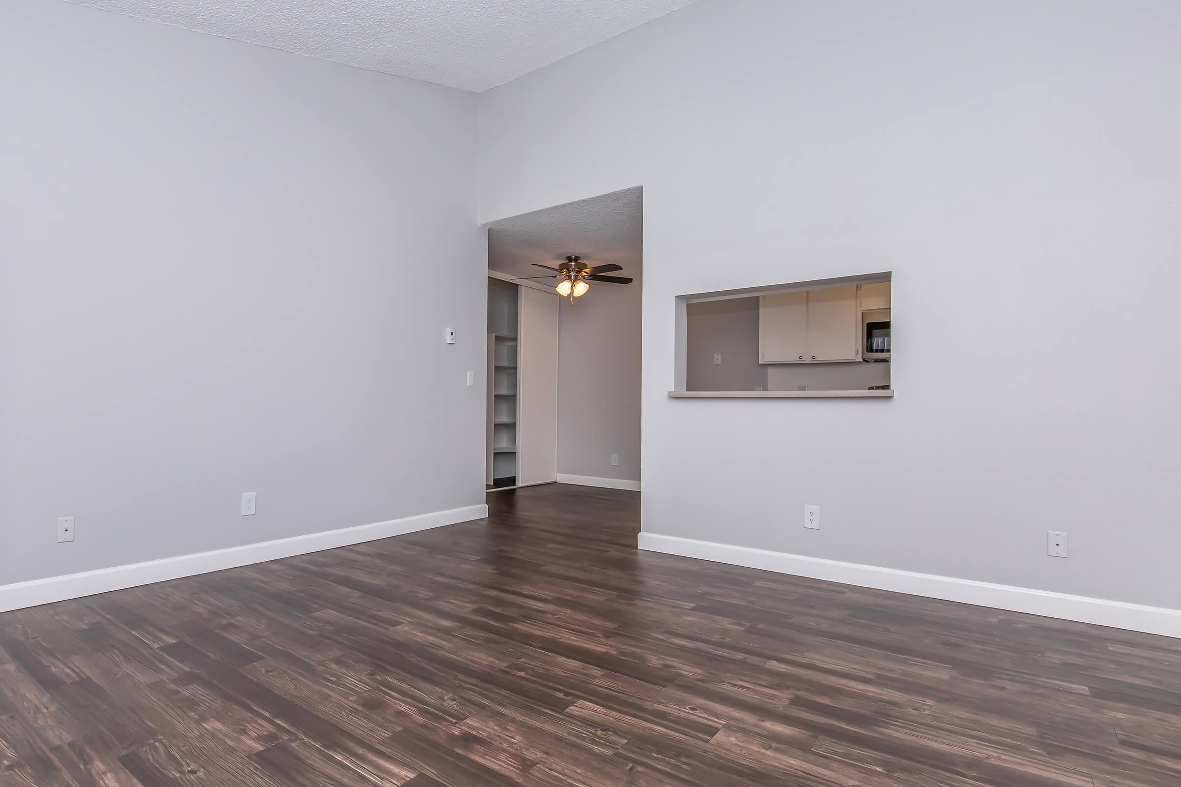 A spacious, empty living area with laminate flooring and light gray walls. There is an opening leading to a kitchen with white cabinets visible in the background. A ceiling fan is located in the corner, and the overall ambiance is bright and uncluttered, suggesting a clean and modern design.