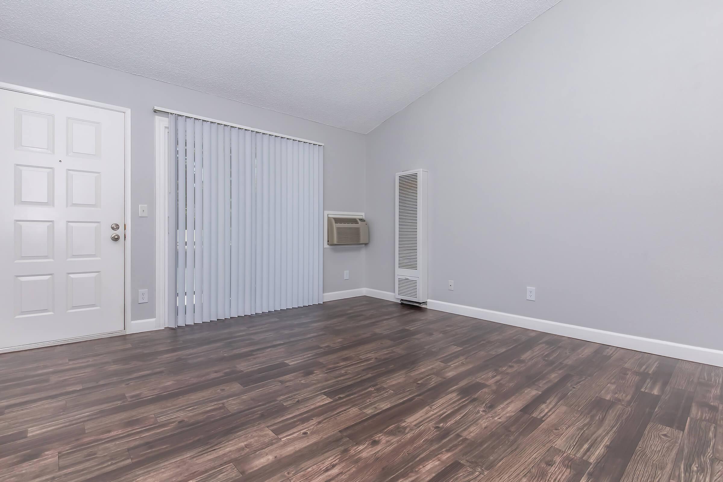 Empty interior of a room featuring light gray walls, a door with a window, vertical blinds, an air conditioning unit, and laminate flooring. The space has a minimalistic design with no furniture or decorations, emphasizing the clean and open feel of the area.