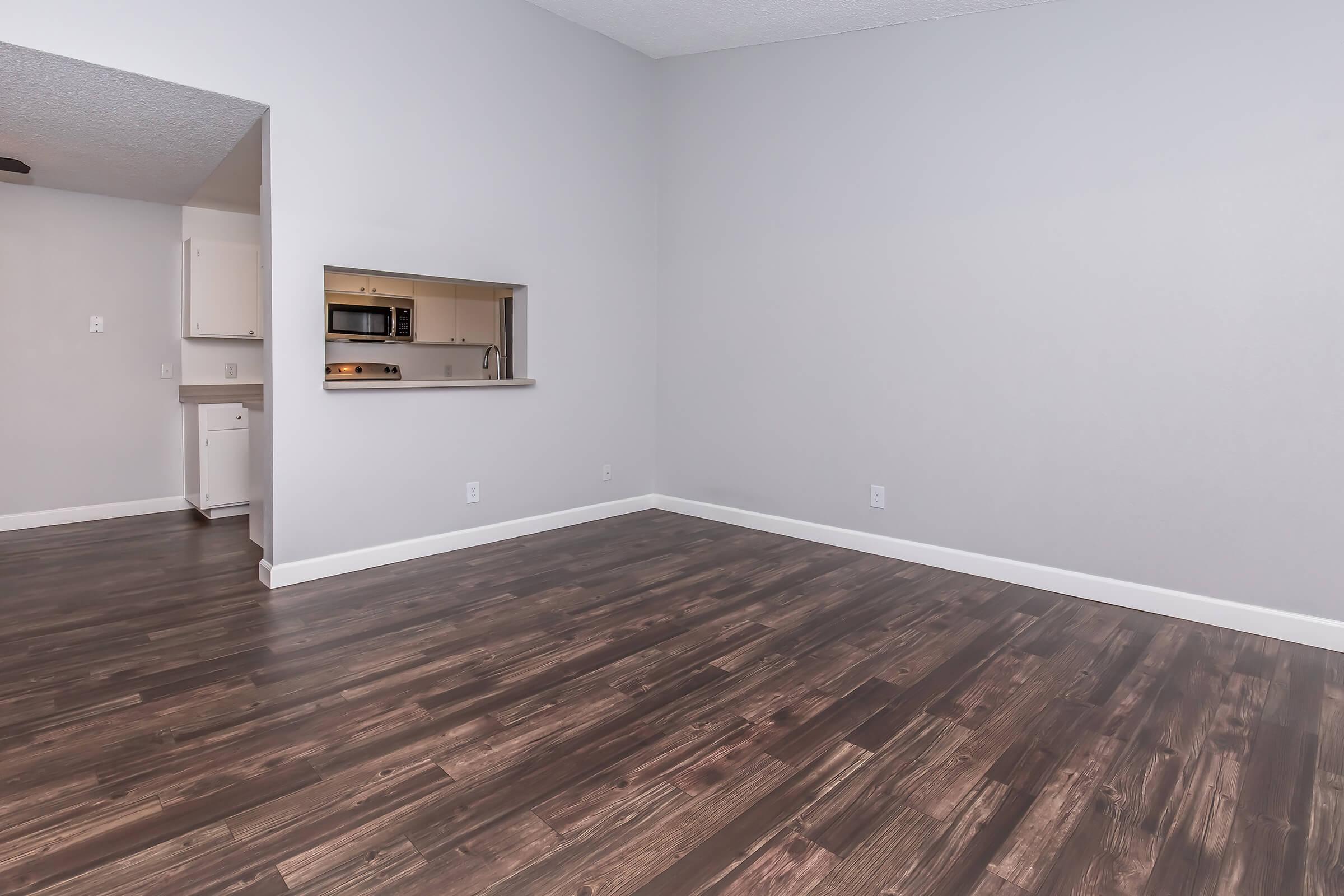 Open living space with light gray walls and wooden flooring. A kitchen is partially visible through an open pass-through counter. The area is well-lit, featuring a high ceiling, with no furniture present, creating a spacious and minimalist feel.