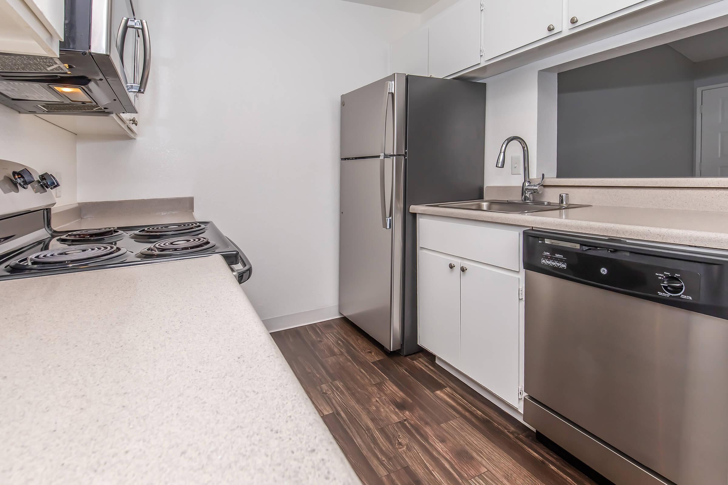 Modern kitchen featuring stainless steel appliances, including a refrigerator, dishwasher, and electric stove. Counters are light-colored with a smooth finish, and cabinetry is white. The flooring is dark wood, creating a contrast with the lighter elements of the kitchen.