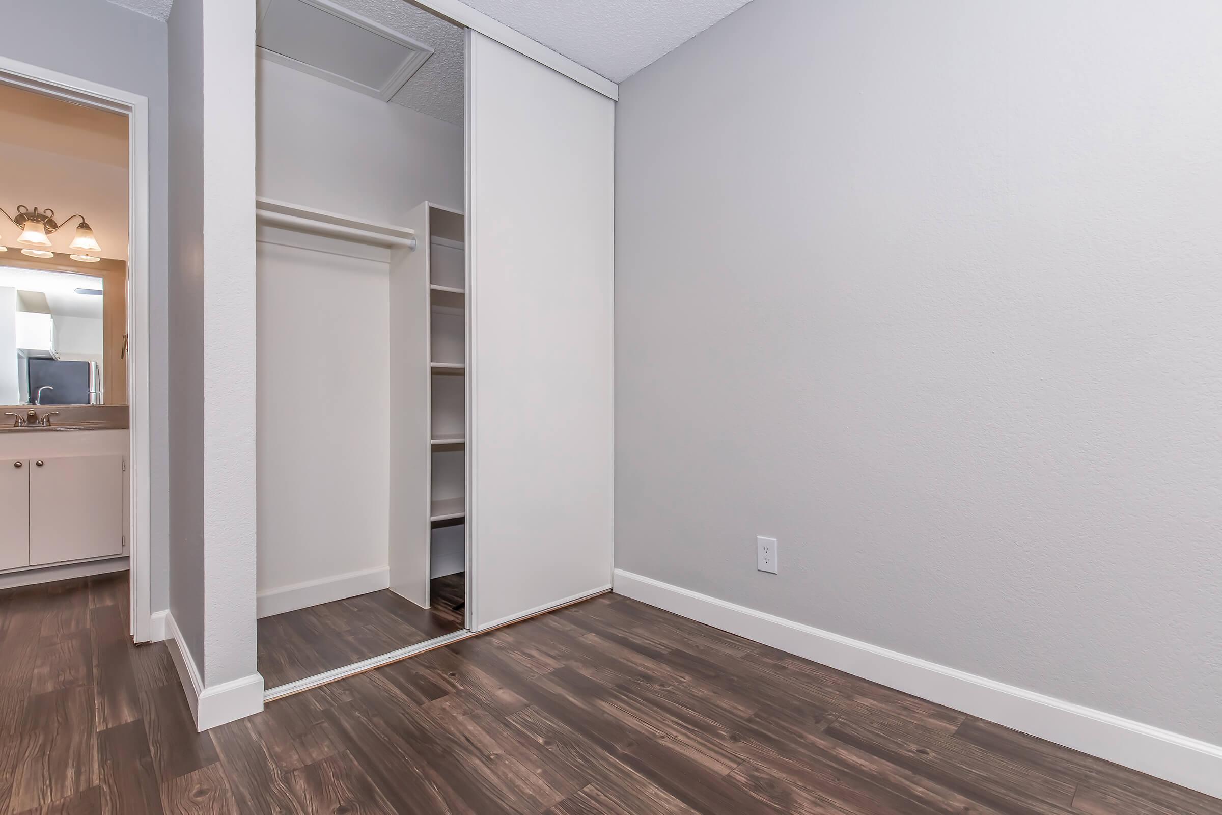 A room with light gray walls and a large mirror closet. The floor has a dark wood finish. In the background, a glimpse of a bathroom can be seen with a light fixture and a vanity. The space appears clean and modern, suitable for a bedroom or dressing area.
