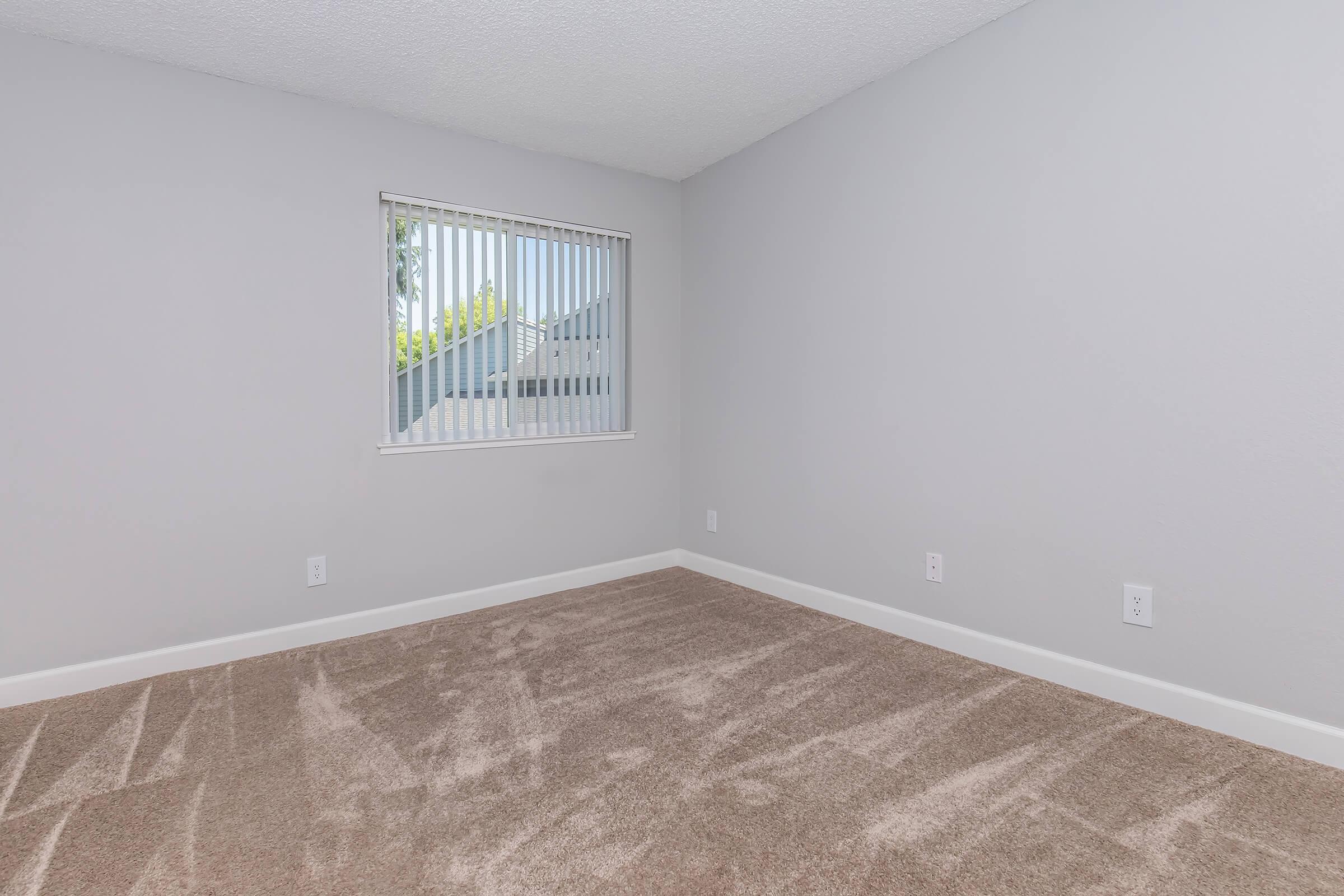 A vacant room with light gray walls and carpeted flooring. There's a window with vertical blinds allowing natural light to enter, and the space is minimalist, featuring no furniture or decorations. The overall atmosphere is clean and ready for personal items to be added.