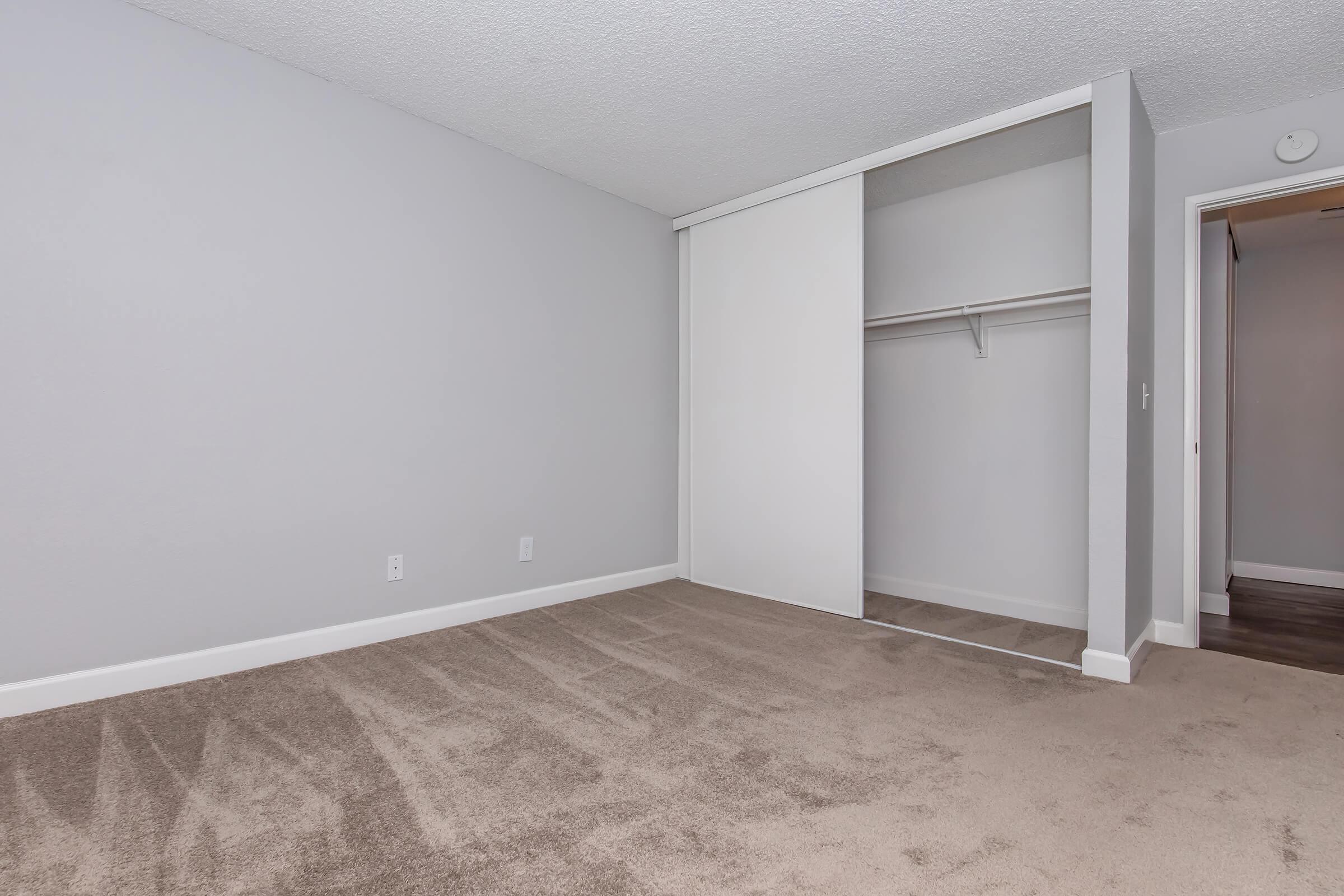 Empty room featuring light gray walls and carpeted flooring. On one side, there is a sliding closet door with a mirror. The room has minimal furnishings and a simple, clean design, providing a blank canvas for potential decoration. A doorway is visible in the background.
