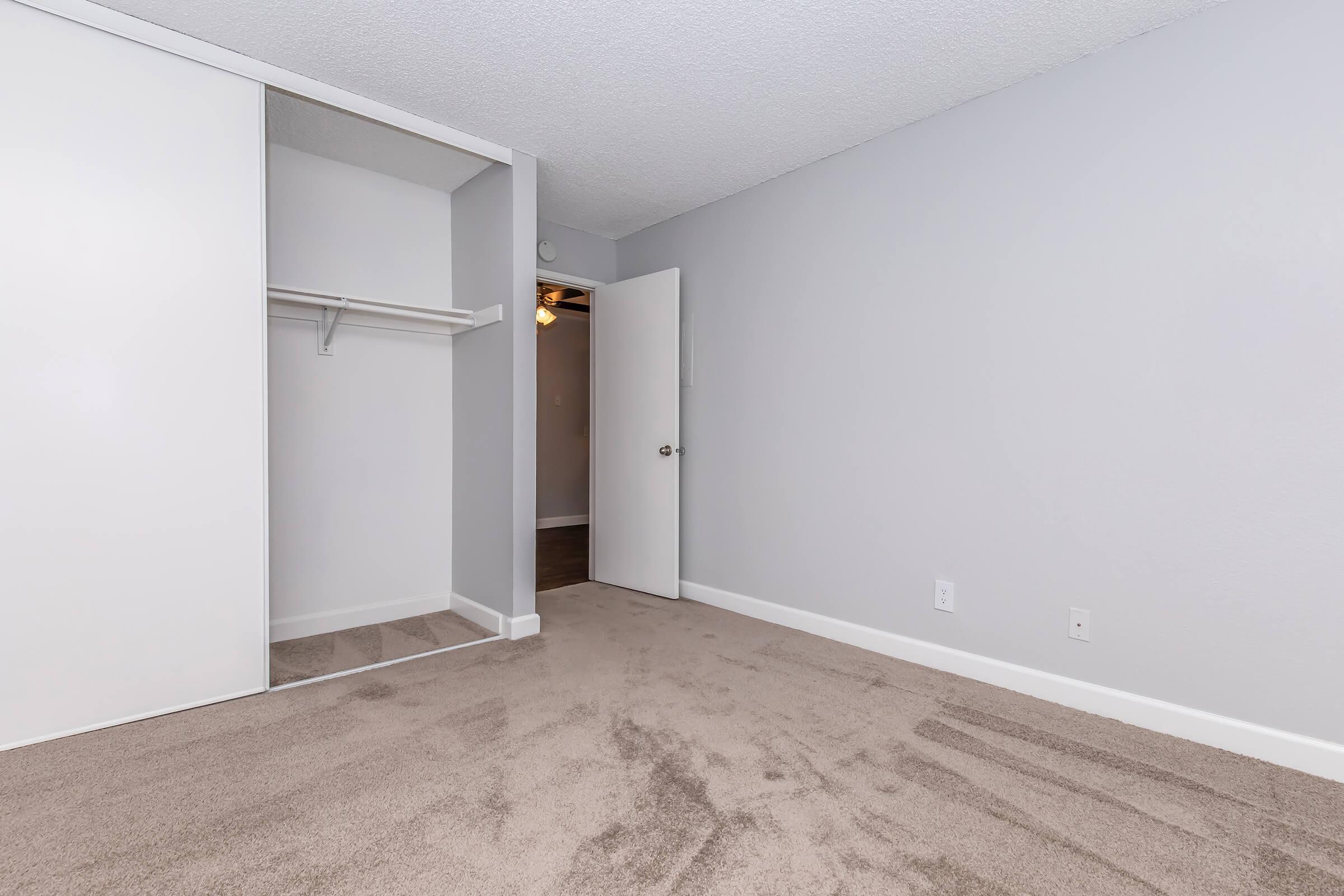 A clean, empty room with freshly painted gray walls and light carpet. A white closet with a hanging rod is visible on the left, and a partially open door leads to another space. The room is well-lit, creating a spacious and inviting atmosphere.