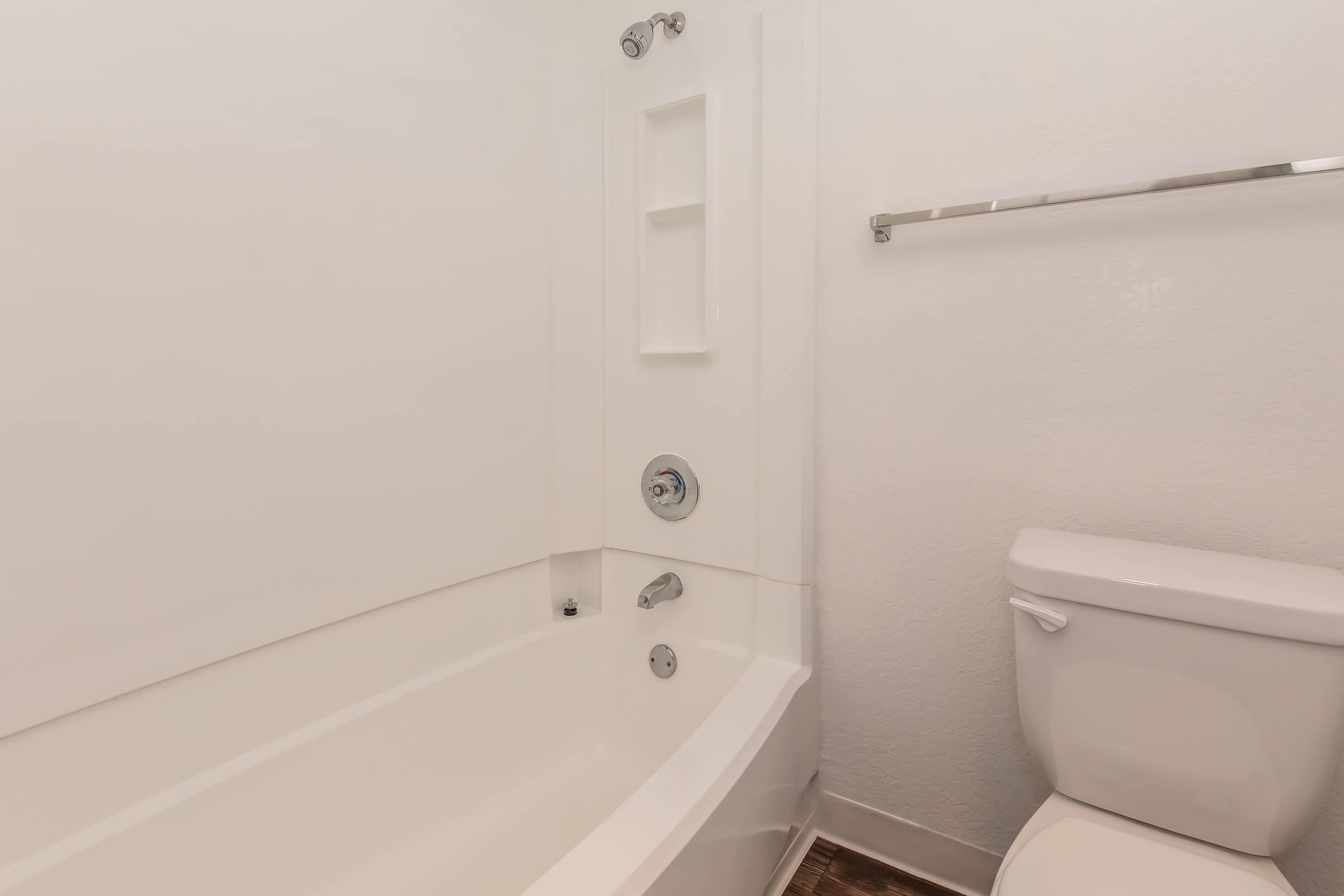 A clean, minimalistic bathroom featuring a white bathtub with a built-in shelf, a chrome faucet, and a sleek toilet. The walls are painted white, and the flooring is a warm wood tone, creating a simple and modern aesthetic.