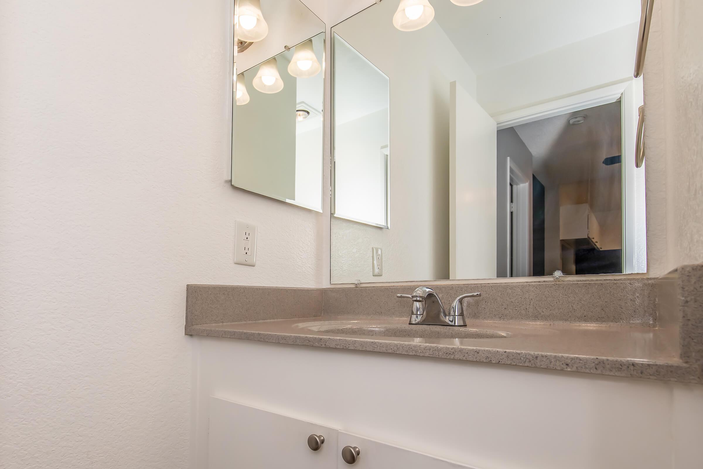 A clean bathroom vanity with a double sink, modern chrome faucets, and a light gray countertop. The wall features two mirrors above the sinks. The background shows a doorway leading to another room and light-colored walls, creating a bright and spacious atmosphere.